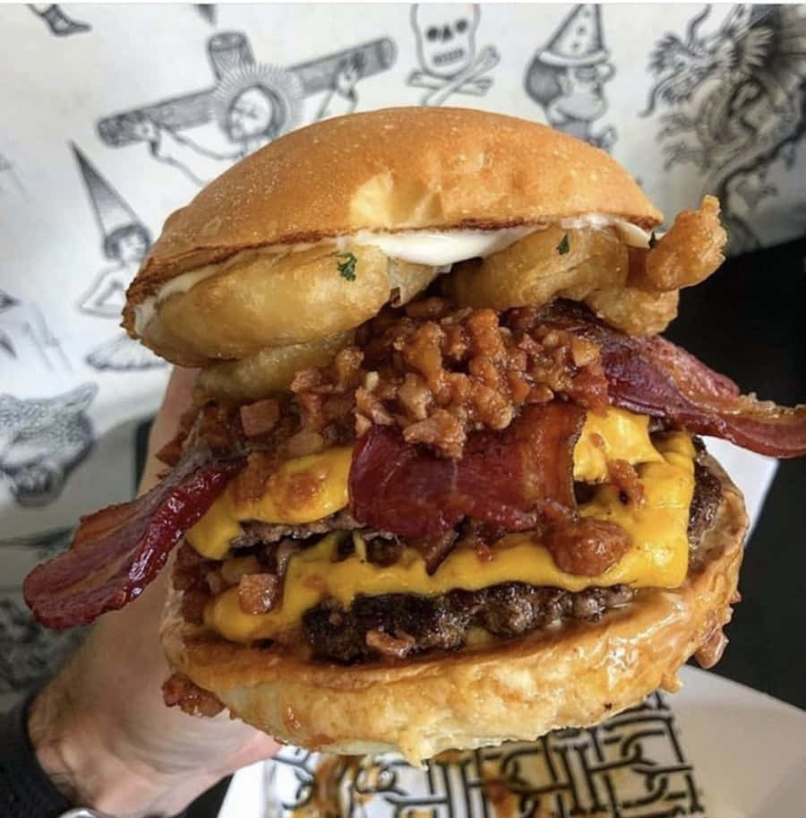fast food 桌布 壁紙 — 一隻手拿著一個高聳的漢堡，上面有脆脆的培根、融化的起司、焦糖洋蔥和炸洋蔥圈，背景是圖案。