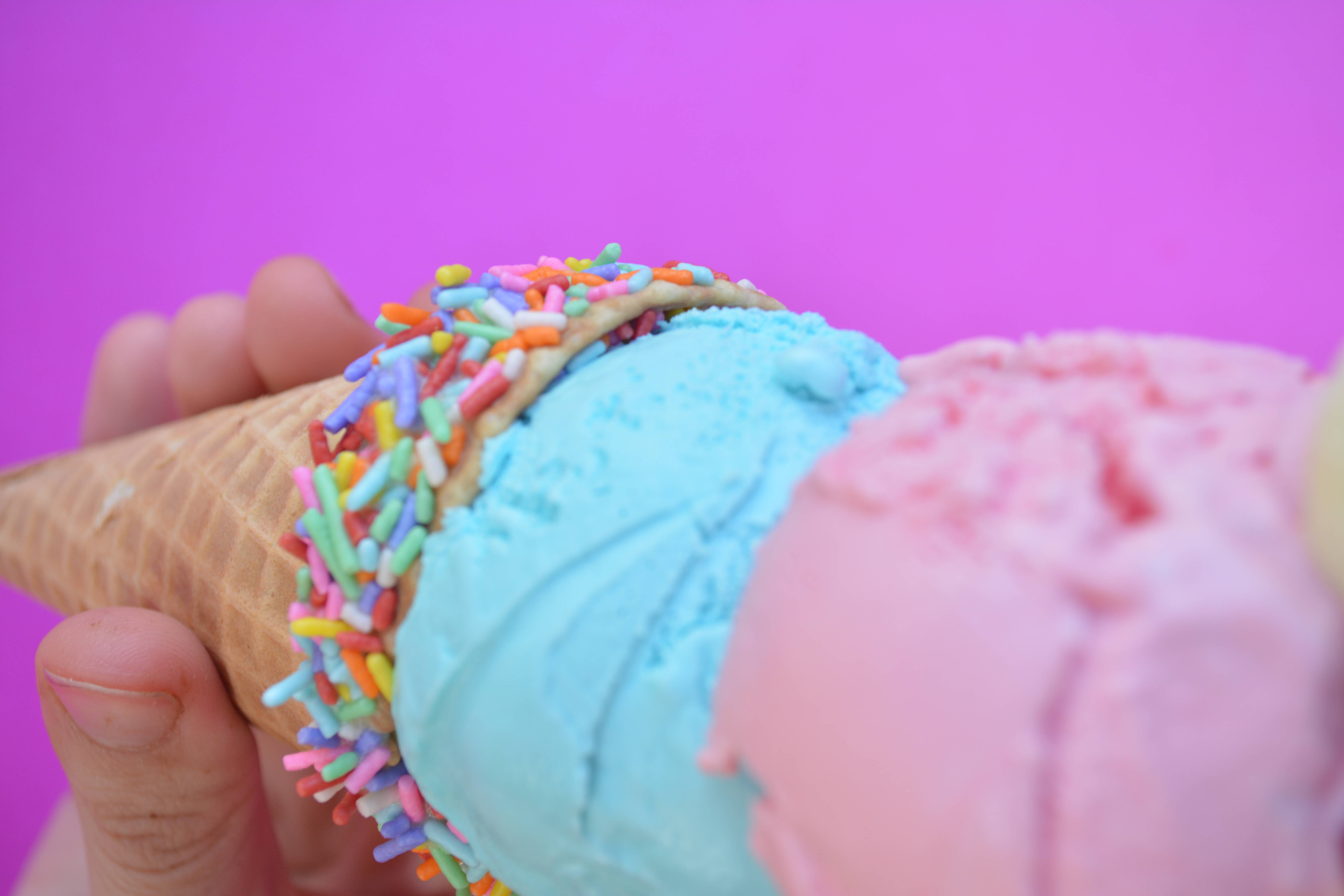 Ice Cream 桌布 壁紙 — 一隻手拿著一個鬆餅筒，上面有藍色和粉紅色的冰淇淋，綴以色彩繽紛的糖粒，背景是鮮豔的紫色。