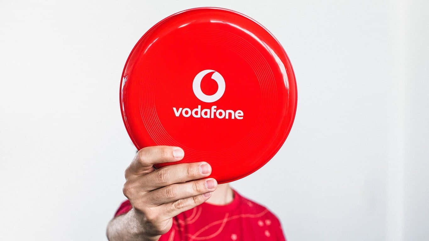 1366x768 Frisbee 桌布 壁紙 — 一隻手拿著一個鮮紅色的Vodafone飛盤，遮住該人的臉，背景為純白色。
