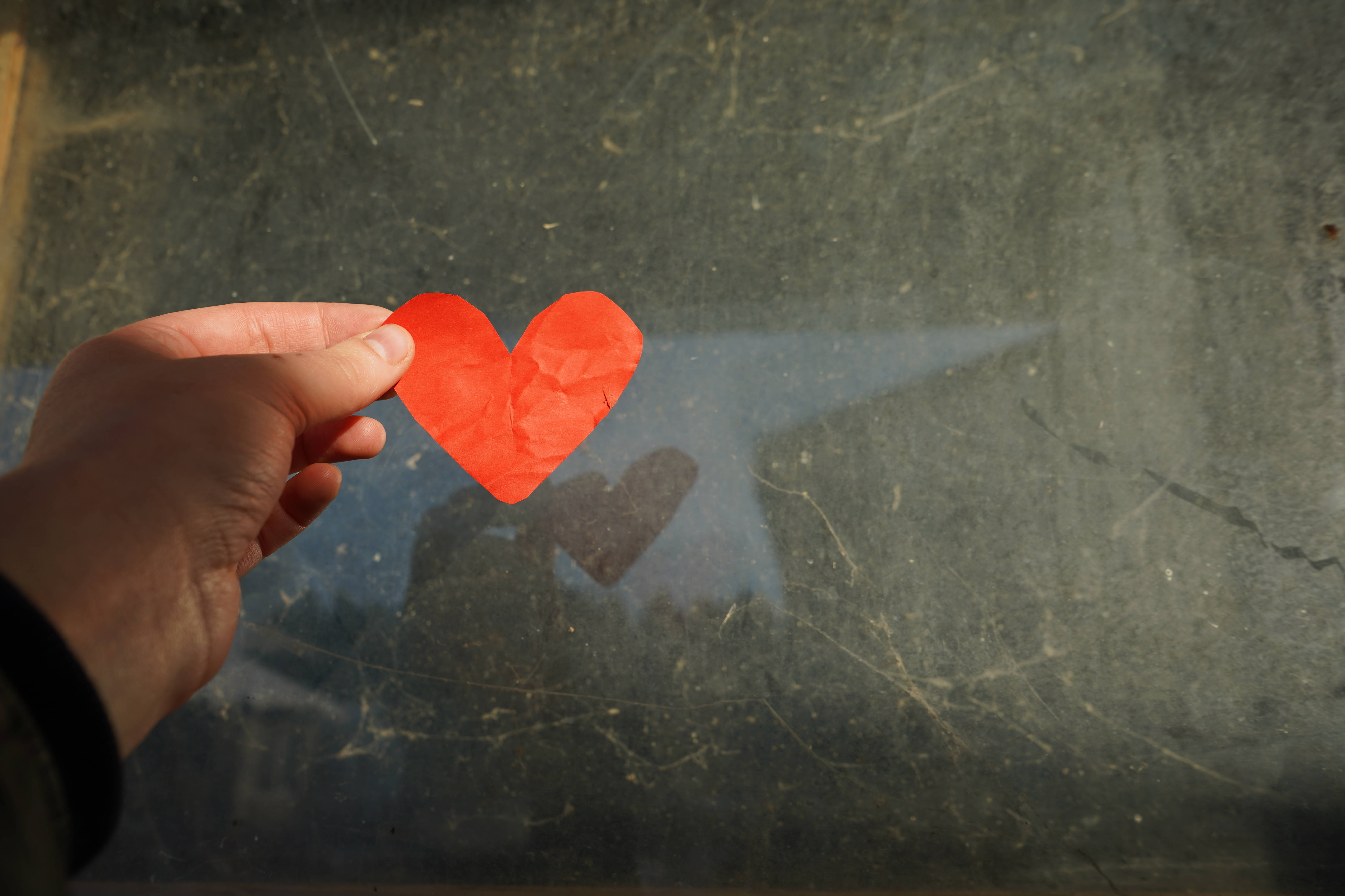 Cute Heart 桌布 壁紙 — 一隻手拿著一個鮮紅色的紙愛心，靠近一塊有刮痕的玻璃窗，柔和陽光穿透而過。