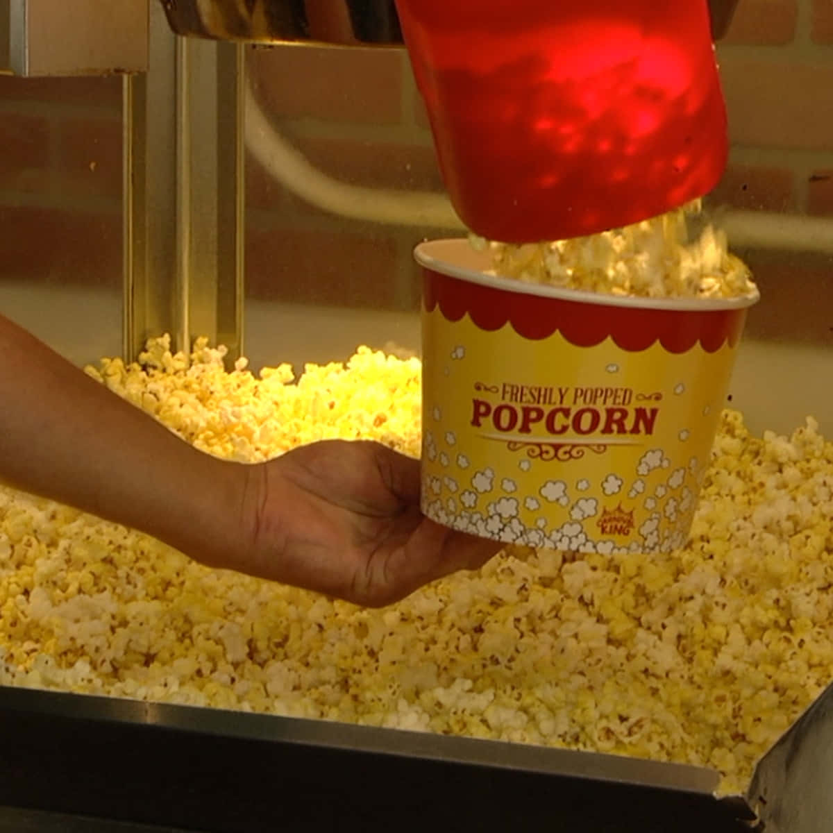 Popcorn 桌布 壁紙 — 一隻手拿著一個黃色爆米花桶，新鮮爆好的爆米花從紅色滑槽倒入桶中，地點是小賣部。
