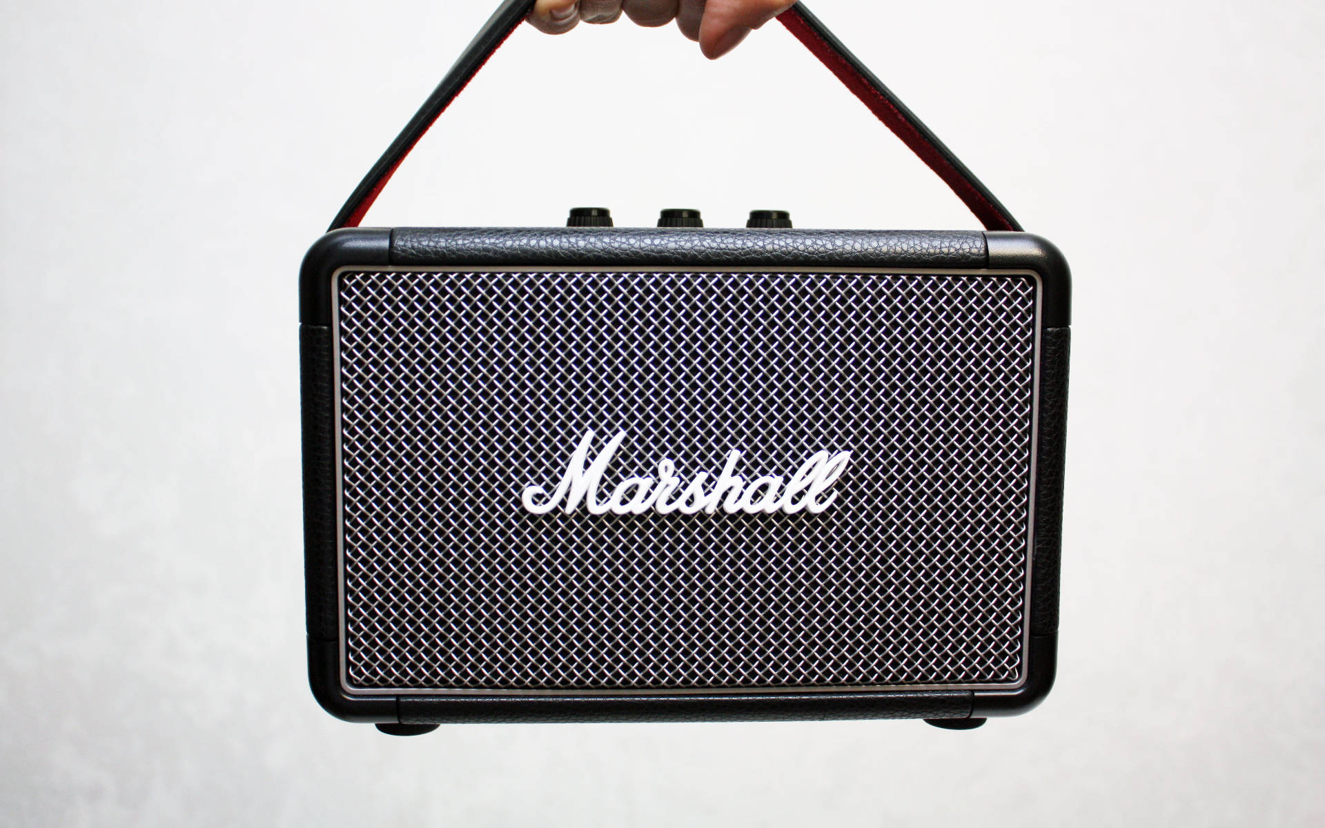Marshall 桌布 壁紙 — 一隻手拿著一個黑色Marshall便攜式揚聲器，帶有紋理格柵和紅色飾邊手柄，背景為純白色。