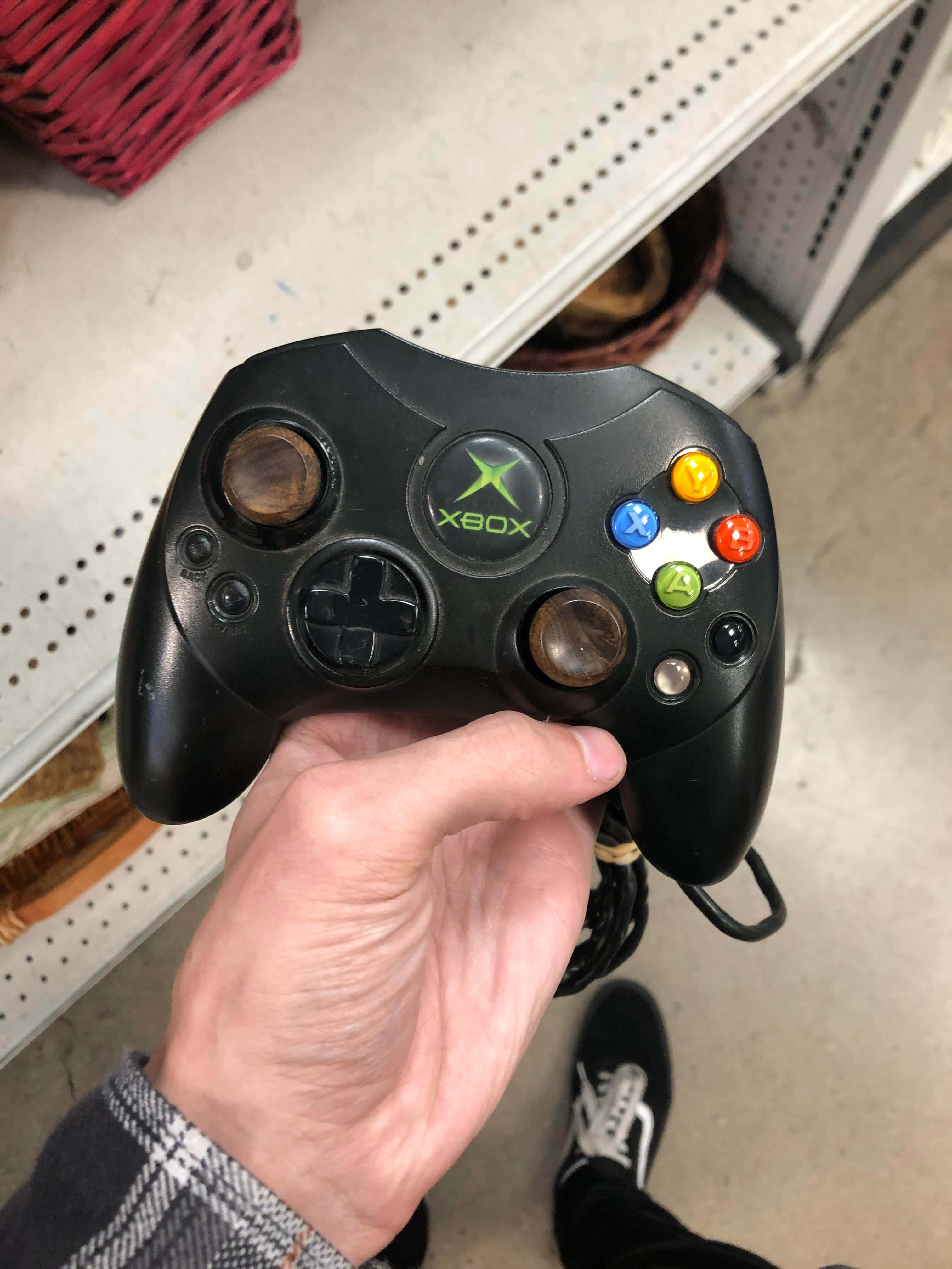 Xbox 桌布 壁紙 — 一隻手拿著一個黑色Xbox 360控制器，其類比搖桿已磨損，背景為穿孔架子。