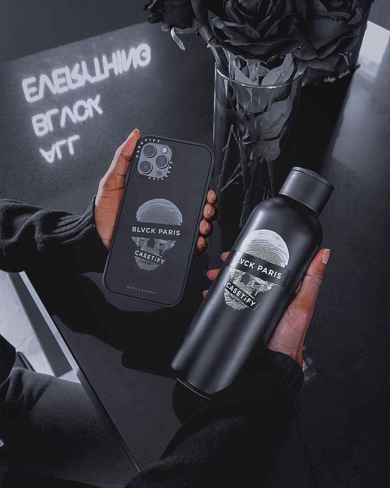 Blvck Paris 桌布 壁紙 — 一隻手拿著一個黑色手機殼和一個瓶子，兩者均帶有BLVCK PARIS品牌標誌，背景為黑色玫瑰和發光文字的暗色背景。