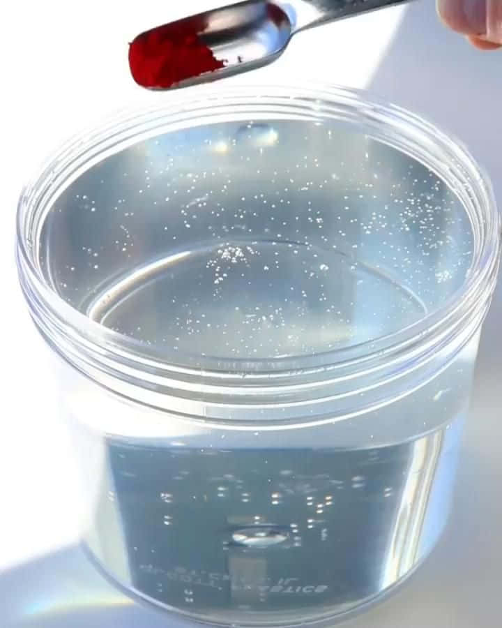 slime 桌布 壁紙 — 一隻手拿著一匙鮮紅色粉末，懸在一個裝有冒泡液體的透明罐子上方。