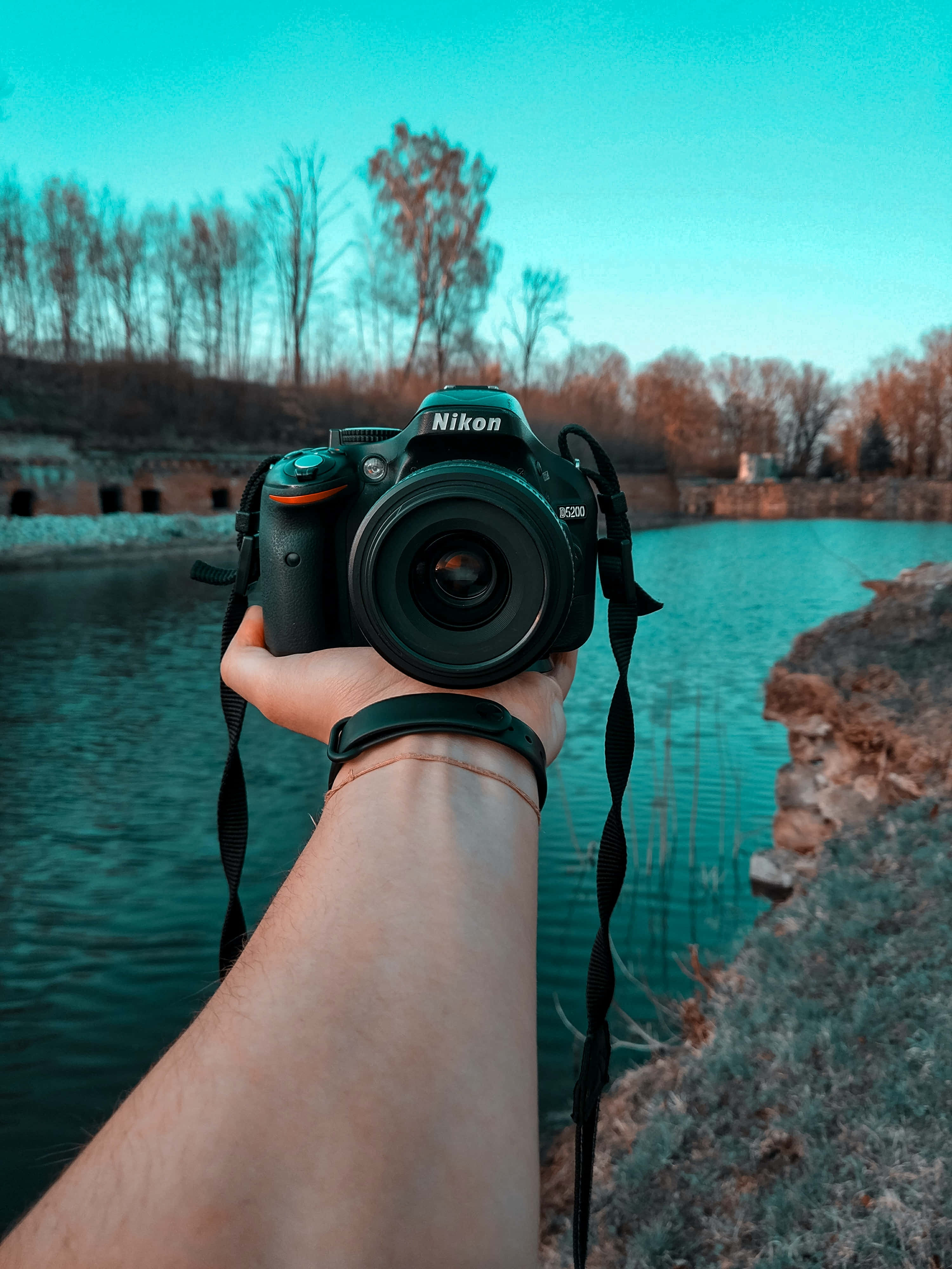 Dslr 桌布 壁紙 — 一隻手拿著一台Nikon DSLR相機朝前，背景是一個藍綠色的湖和光禿禿的樹。