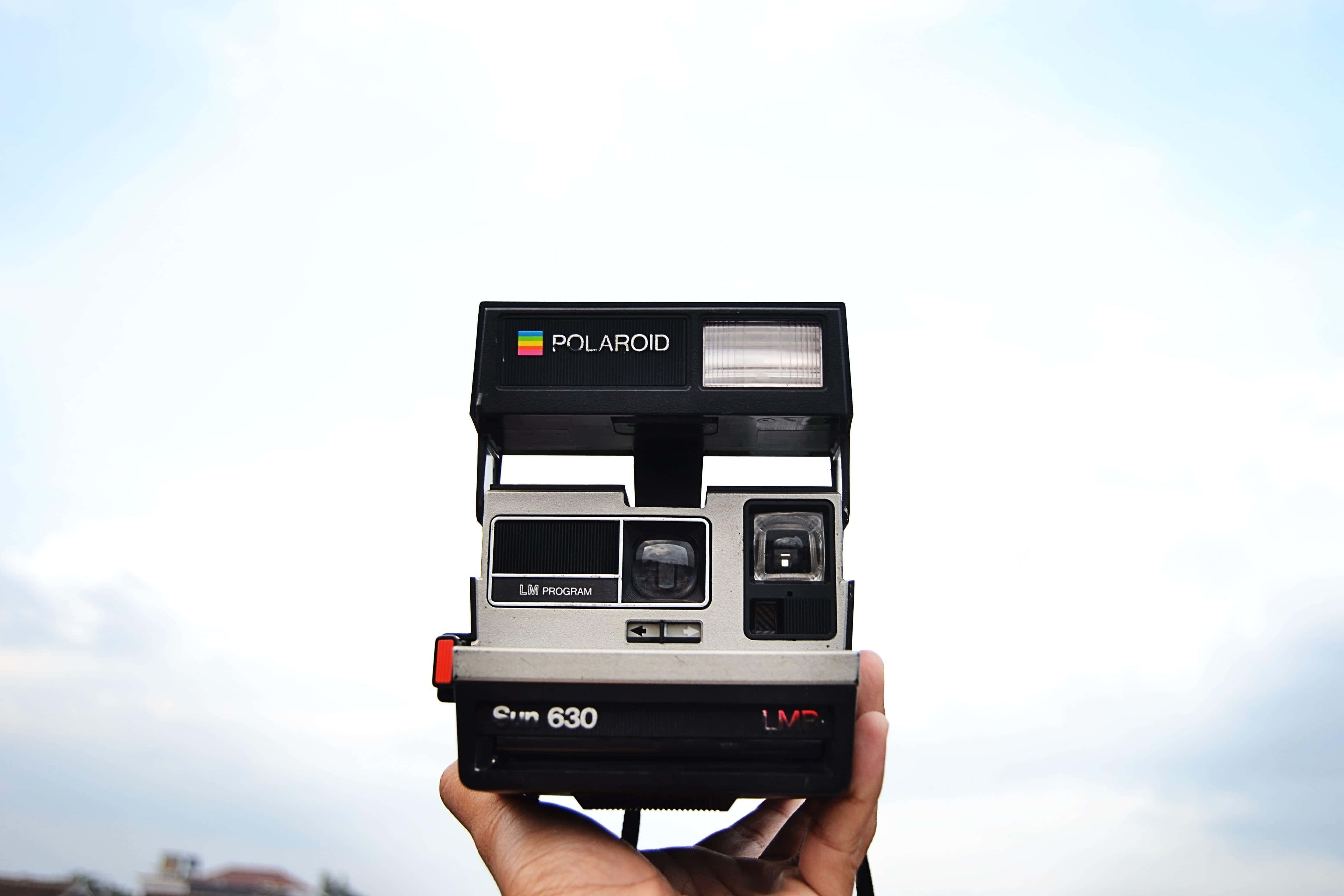 拍立得 桌布 壁紙 — 一隻手拿著一台復古Polaroid Sun 630相機，背景是柔和的雲天。