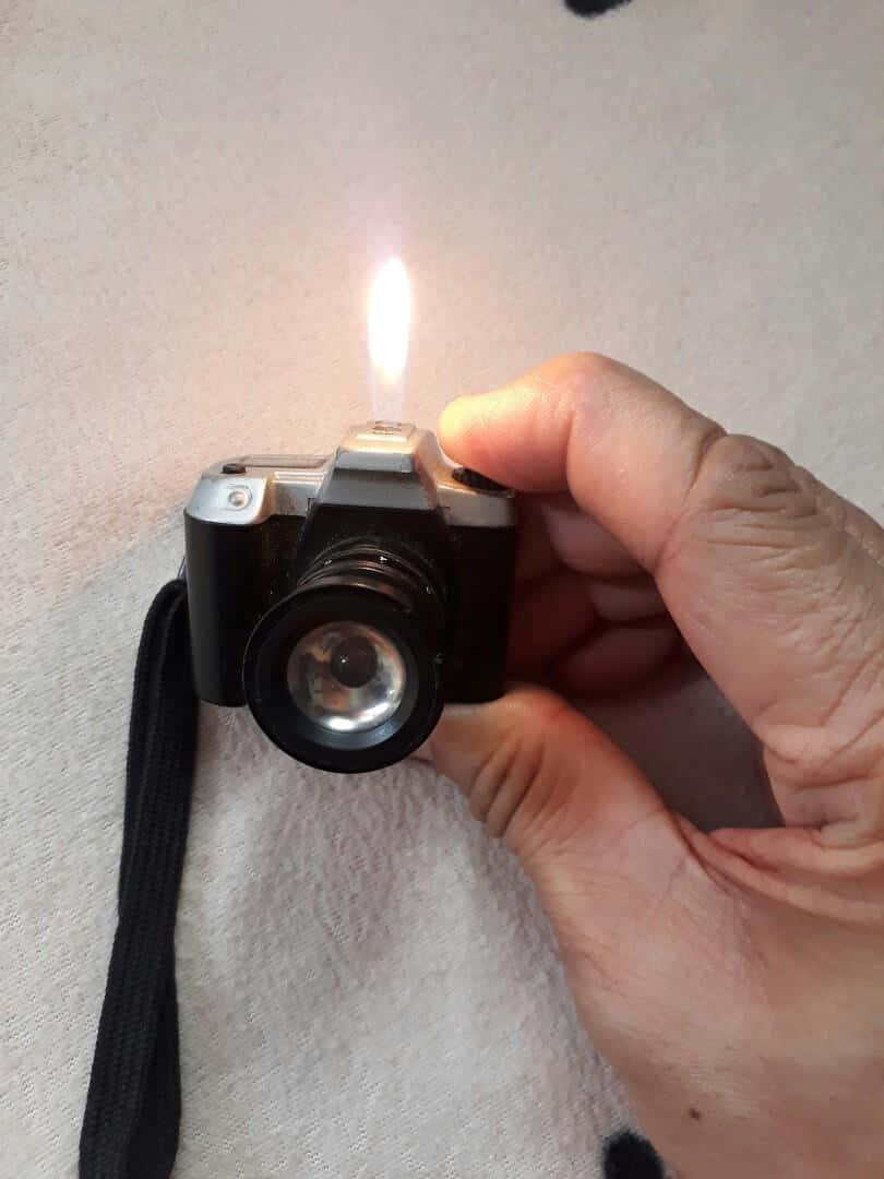 lighter 桌布 壁紙 — 一隻手拿著一台復古相機，頂部噴出明亮的火焰，背景是質感白色布料。