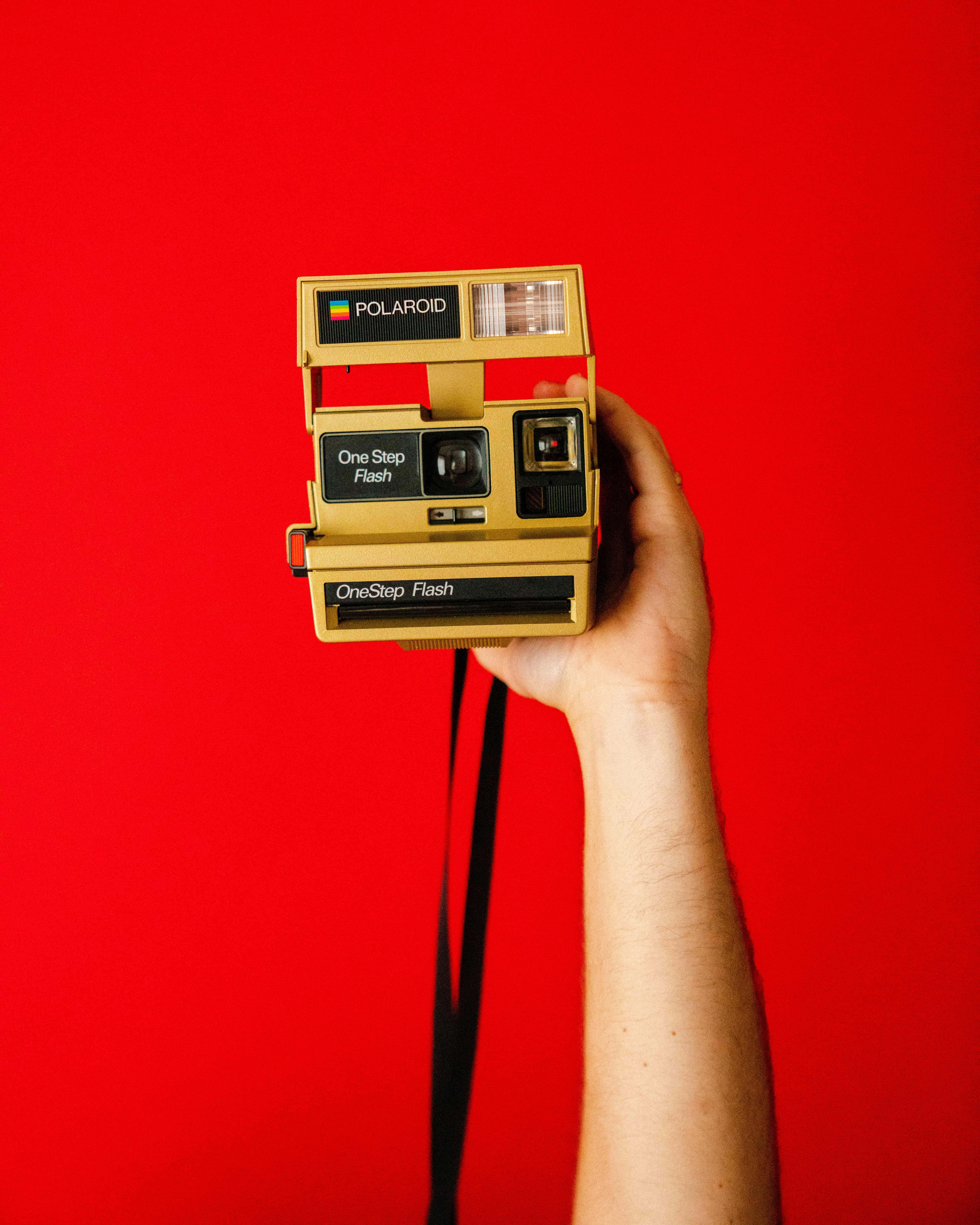 攝影 桌布 壁紙 — 一隻手拿著一台復古金色Polaroid OneStep Flash相機，背景為大膽的紅色。