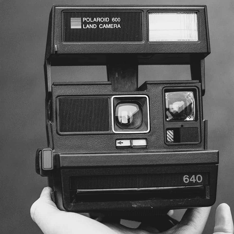 相機 桌布 壁紙 — 一隻手拿著一台復古黑色Polaroid 600 Land相機，背景是單色的純灰色。