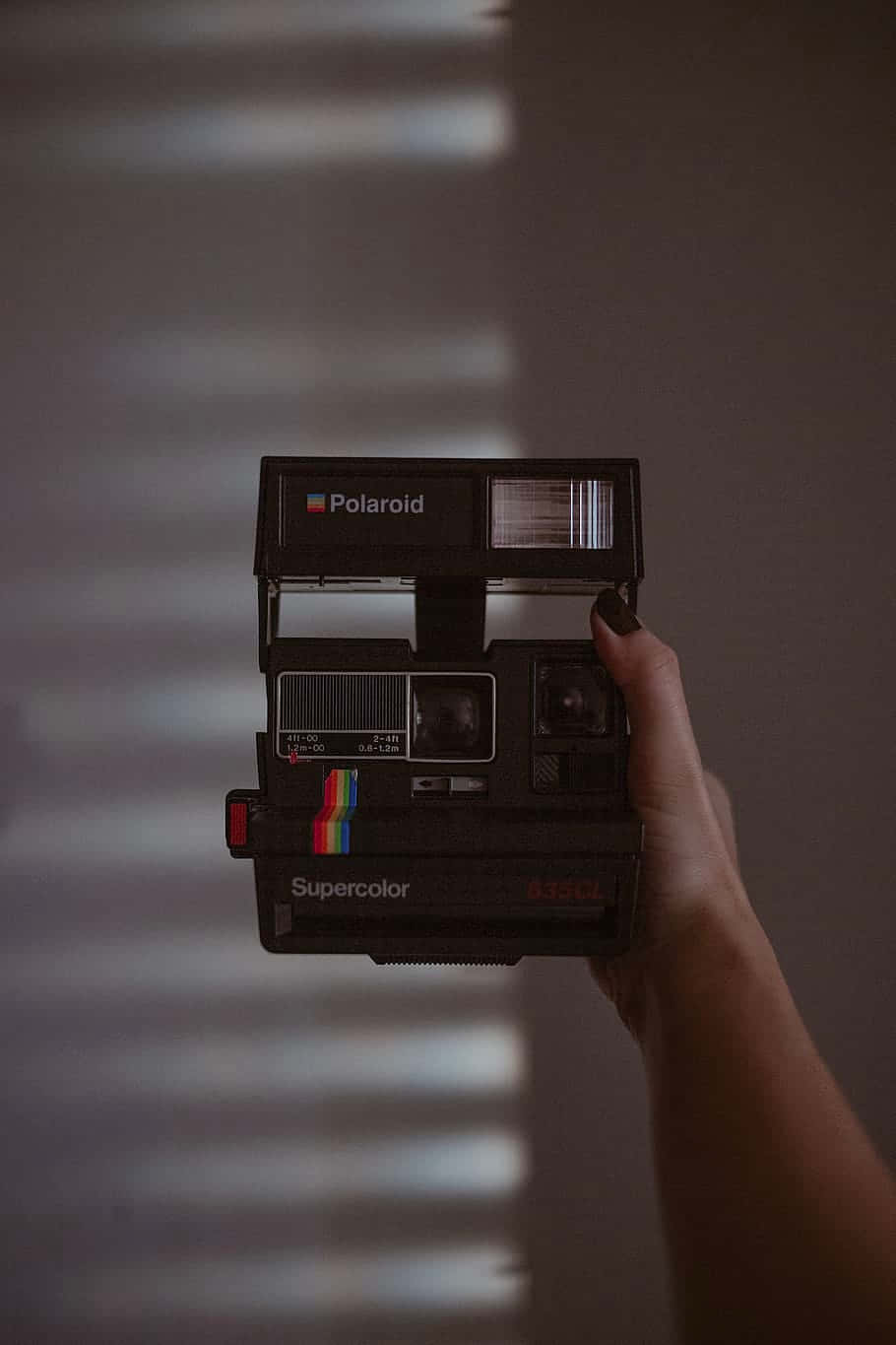 Vintage Camera 桌布 壁紙 — 一隻手拿著一台復古黑色Polaroid Supercolor相機，對著一面有柔和光線和水平光條的牆壁。