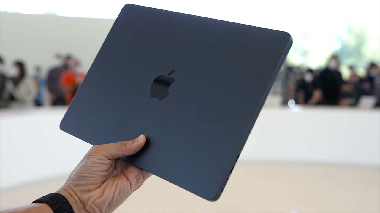 Black Macbook 桌布 壁紙 — 一隻手拿著一台時尚的霧面深藍色MacBook，帶有Apple標誌，背景是模糊的觀眾。