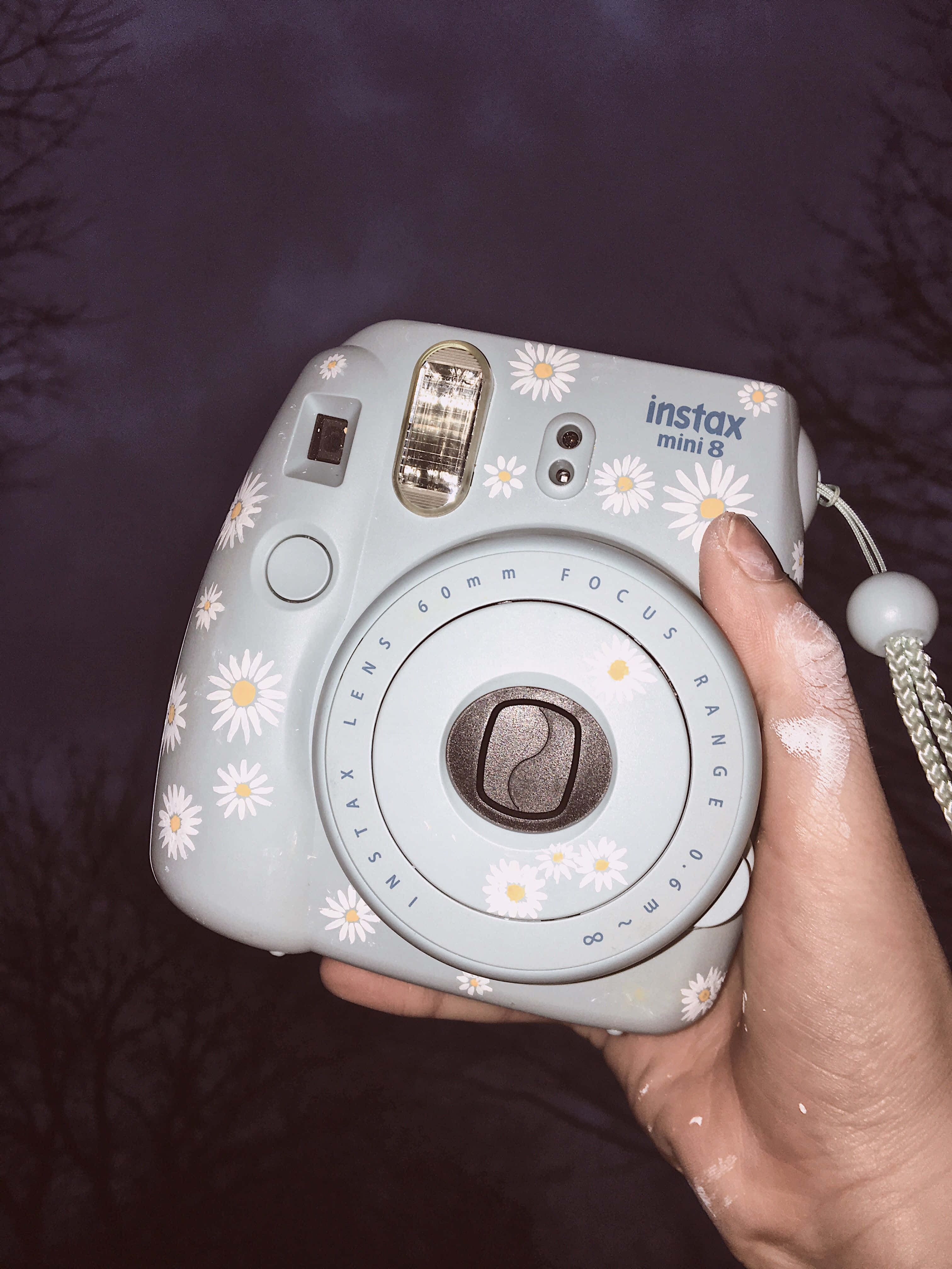 拍立得 桌布 壁紙 — 一隻手拿著一台淺藍色 Instax Mini 8 相機，上面飾有白色雛菊圖案，背景是深紫色的黃昏天空。