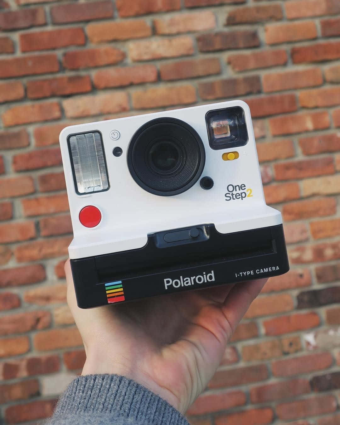 拍立得 桌布 壁紙 — 一隻手拿著一台白色Polaroid One Step 2拍立得相機，背景是溫暖的紅磚牆。