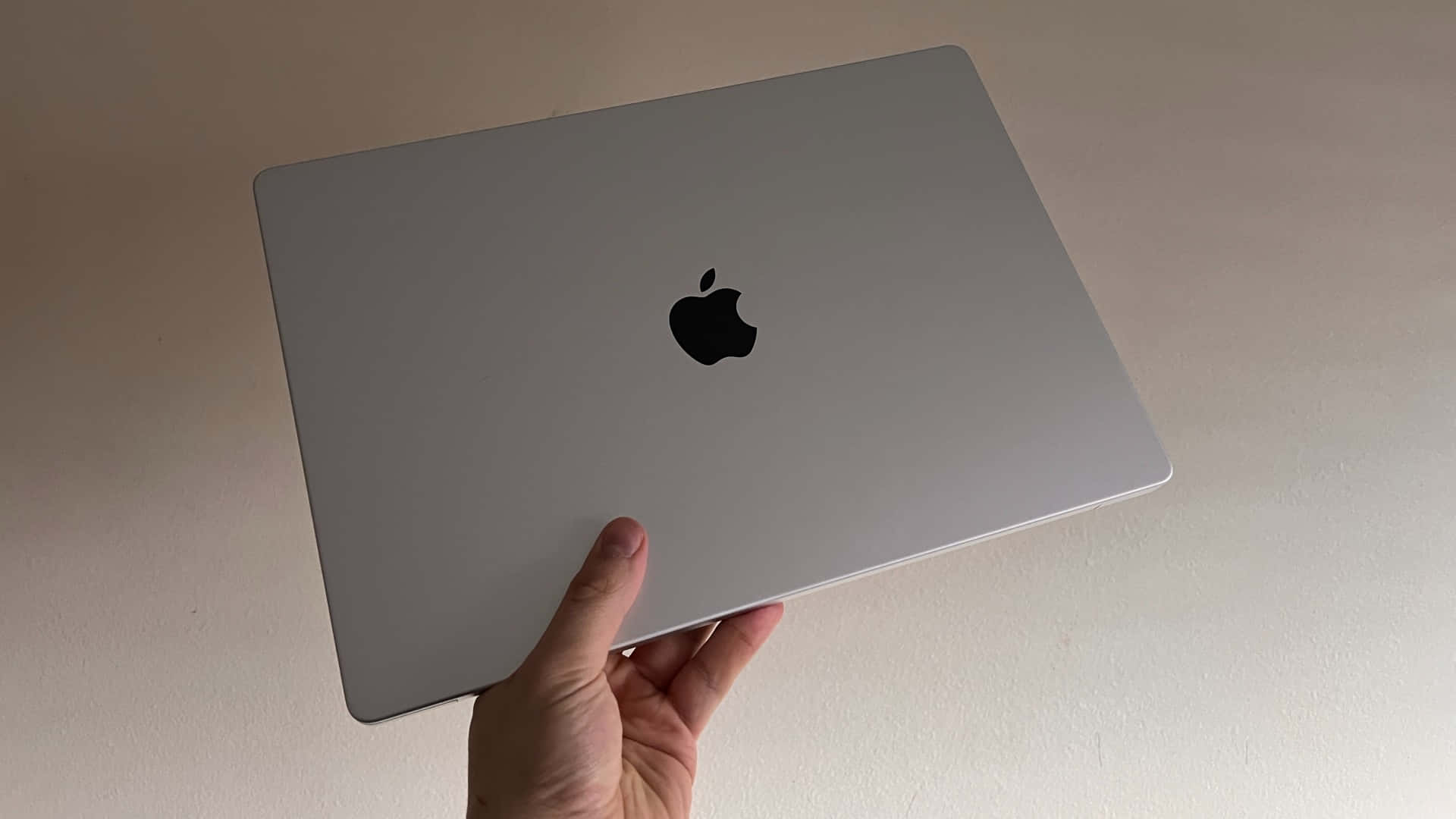 MacBook 動態圖片 桌布 壁紙 — 一隻手拿著一台銀色 MacBook 筆記型電腦，帶有黑色 Apple 標誌，背景是純色米白色牆壁。
