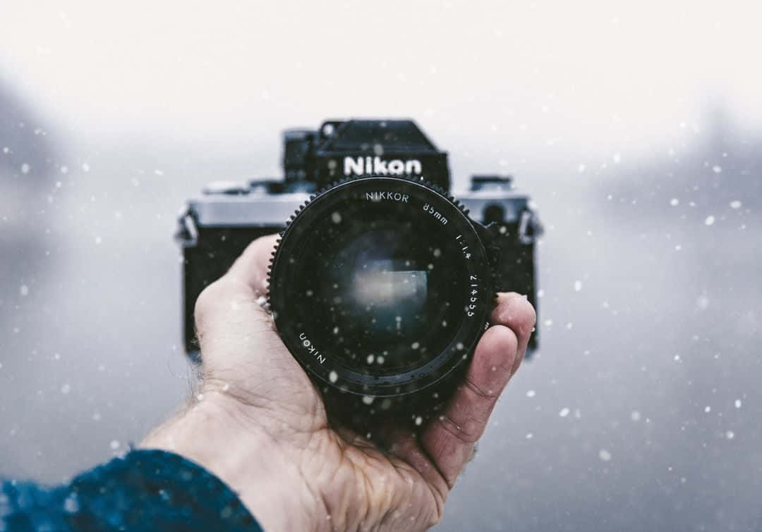 攝影 桌布 壁紙 — 一隻手拿著一台黑色的 Nikon 底片相機，雪花在柔和的灰色冬日天空中飄舞。