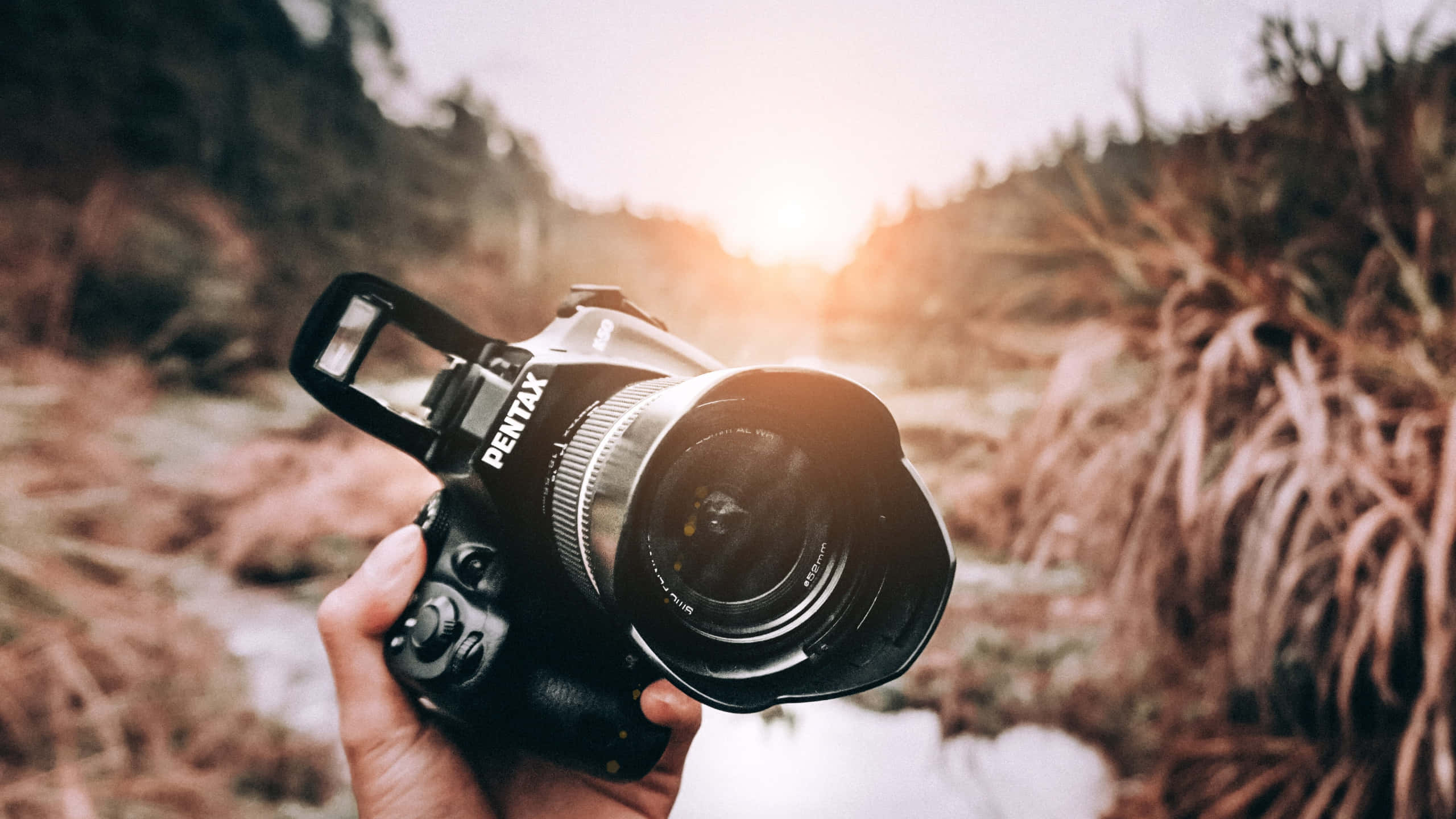 Dslr 桌布 壁紙 — 一隻手拿著一台黑色賓得DSLR相機，背景是陽光普照的戶外，有模糊的草地和山丘。