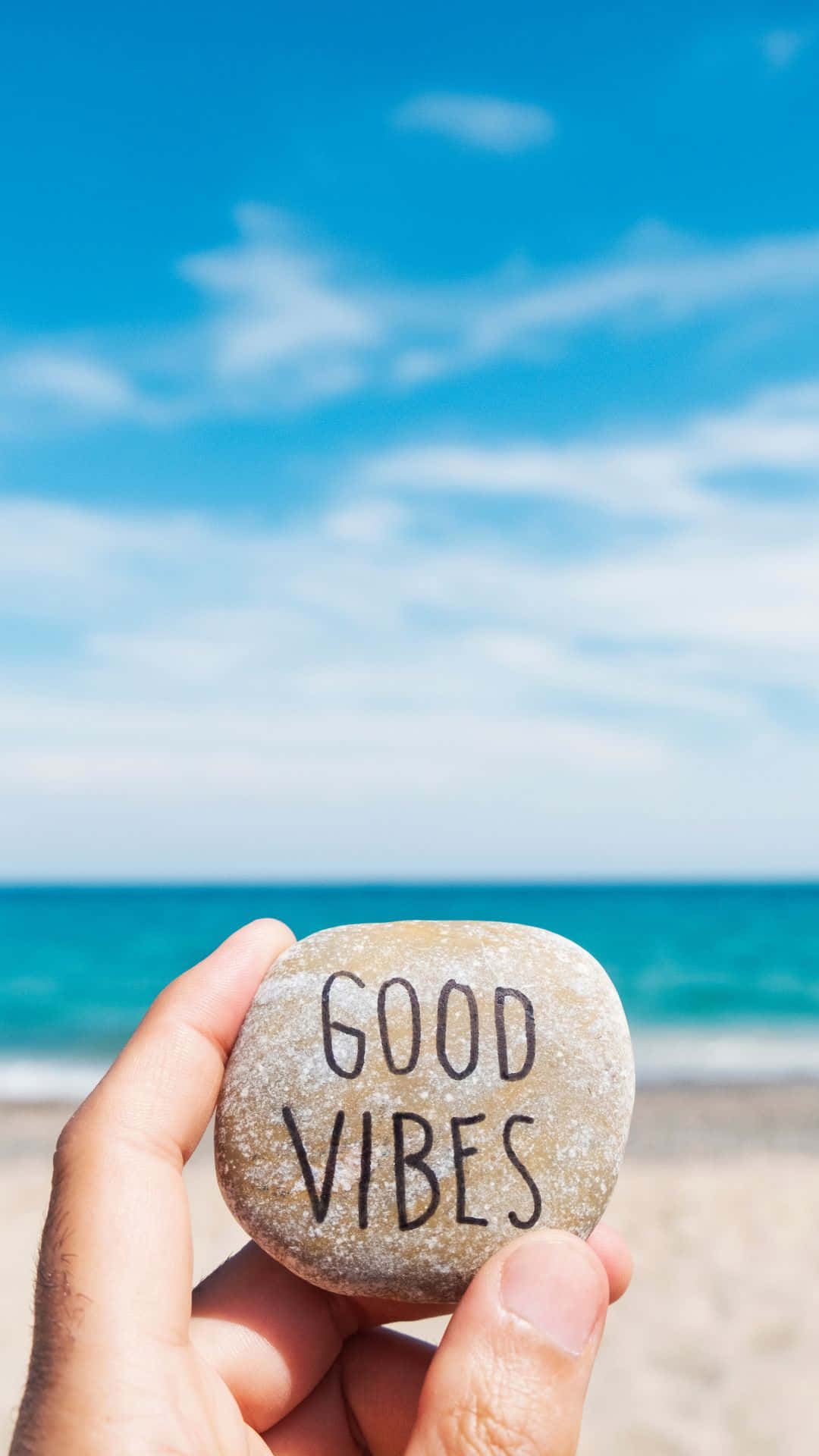 海灘 桌布 壁紙 — 一隻手拿著一塊刻有 'GOOD VIBES' 的光滑石頭，背景是明亮的藍天和碧綠色的海洋。