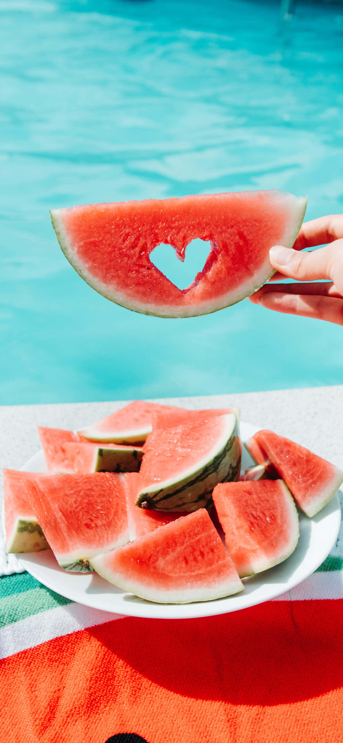 Watermelon iPhone 桌布 壁紙 — 一隻手拿著一塊帶有心形孔的西瓜片，放在一盤西瓜片上方，旁邊是一個藍綠色的游泳池。