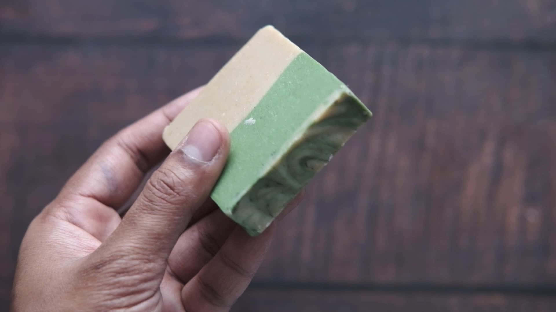 bath 桌布 壁紙 — 一隻手拿著一塊帶有綠色漩渦的雙色肥皂，背景是深色木材。