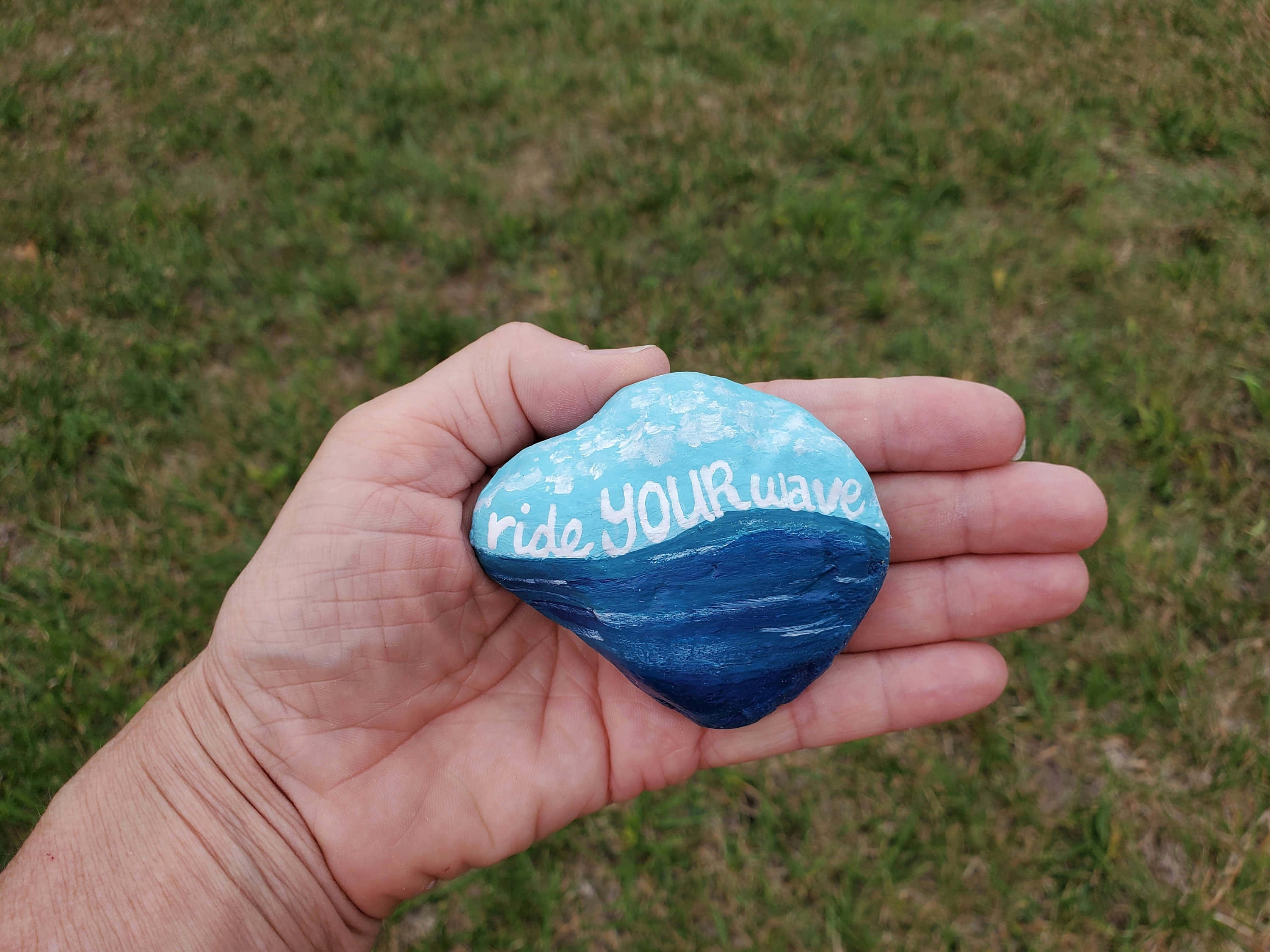 Pebble Art 桌布 壁紙 — 一隻手拿著一塊畫有海浪圖案的石頭，上面用白色草書寫著「ride YOUR wave」，背景是草地。