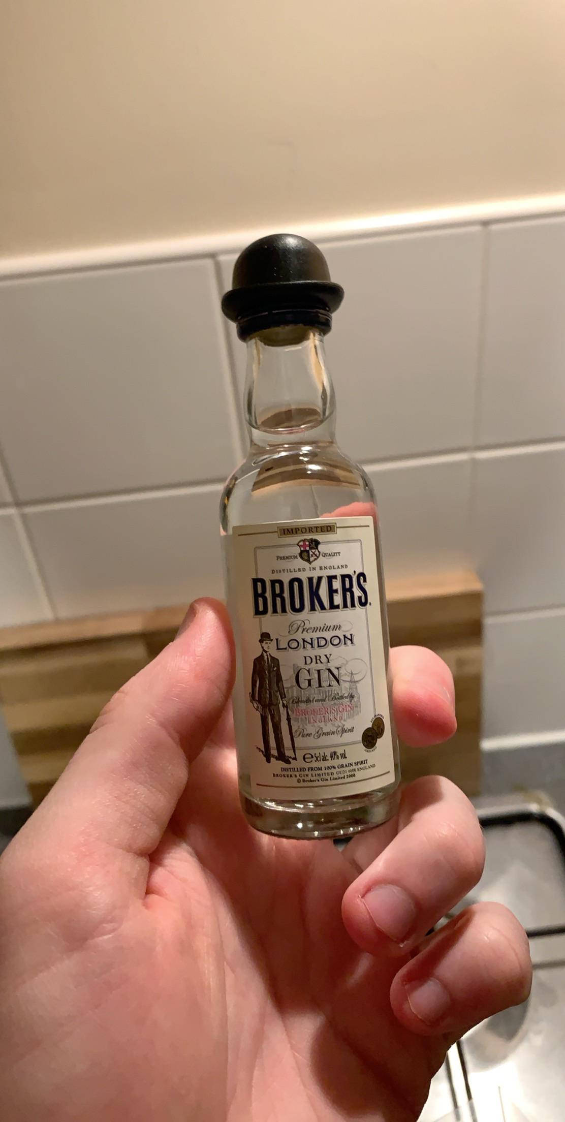 small 桌布 壁紙 — 一隻手拿著一小瓶Brokers Premium London Dry Gin，背景是瓷磚廚房。