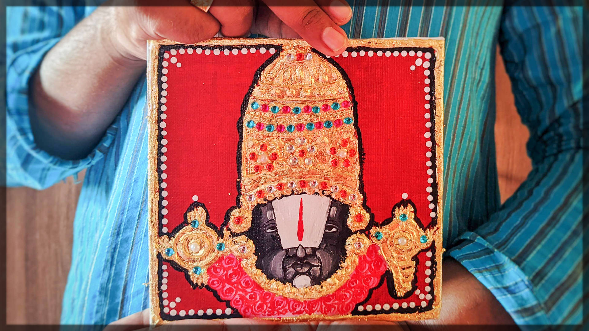 Folk Art 桌布 壁紙 — 一隻手拿著一幅鮮紅色框的藝術品，描繪一位鑲滿珠寶的神祇，戴著金色皇冠和華麗首飾，背景是鮮明的紅色。