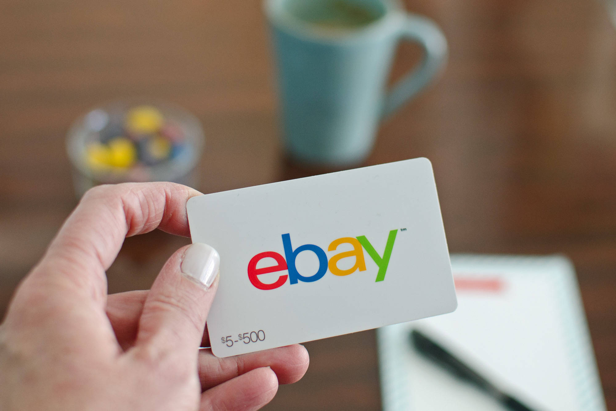eBay 桌布 壁紙 — 一隻手拿著一張價值5到500美元的eBay禮品卡，背景是模糊的木桌，上面有杯子和糖果。