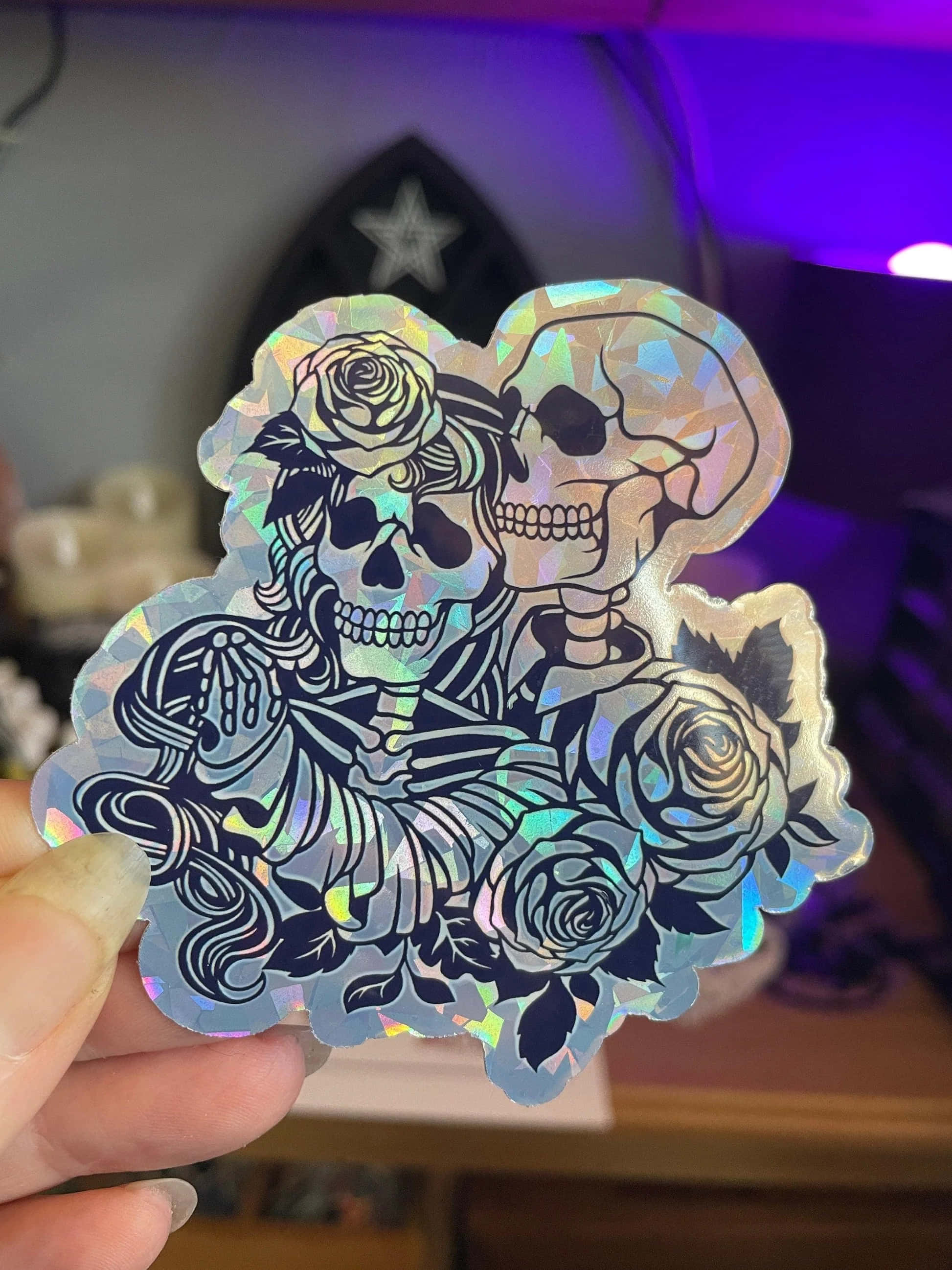 Dark Love 桌布 壁紙 — 一隻手拿著一張全息貼紙，上面有兩個骷髏頭與玫瑰纏繞，在紫色環境光下。
