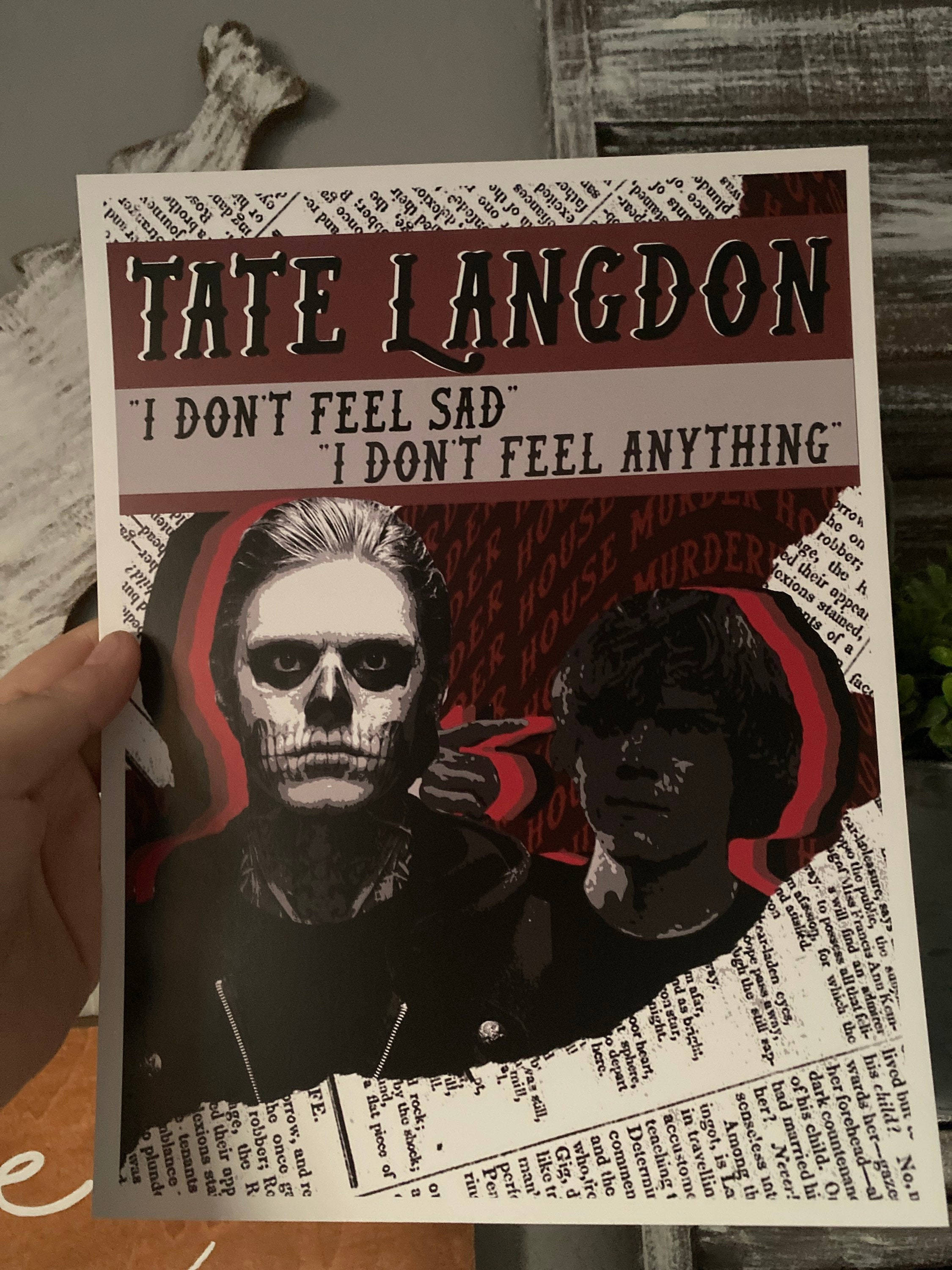 Tate Langdon 桌布 壁紙 — 一隻手拿著一張泰特·蘭登的海報，上面有骷髏臉和文字引言，背景是報紙拼貼。