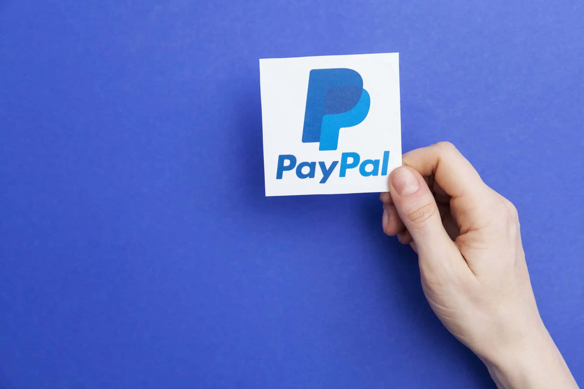 PayPal 桌布 壁紙 — 一隻手拿著一張白色卡片，上面顯示藍色PayPal標誌，背景為純藍色。