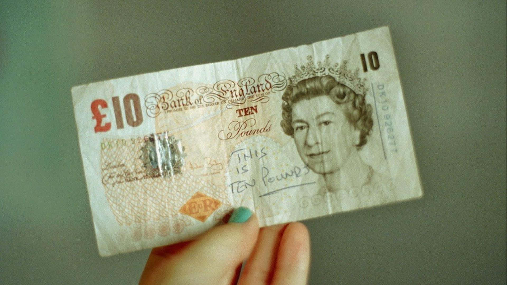 Queen Elizabeth 桌布 壁紙 — 一隻手拿著一張皺巴巴的十英鎊英國紙幣，上面有手寫文字覆蓋在女王肖像上，背景是柔和的灰色。