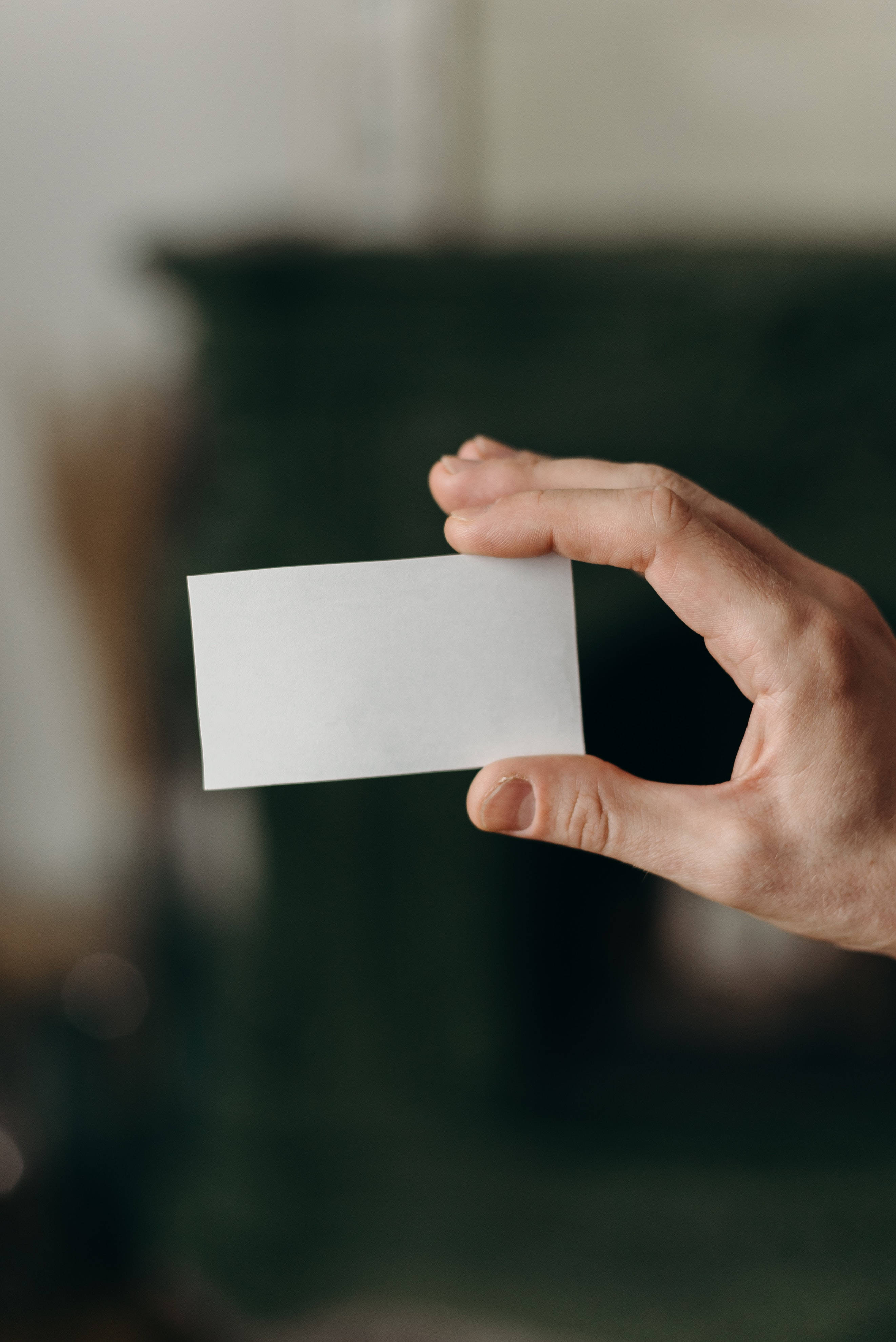 paper 桌布 壁紙 — 一隻手拿著一張空白白色名片，背景是柔和模糊的深綠色。