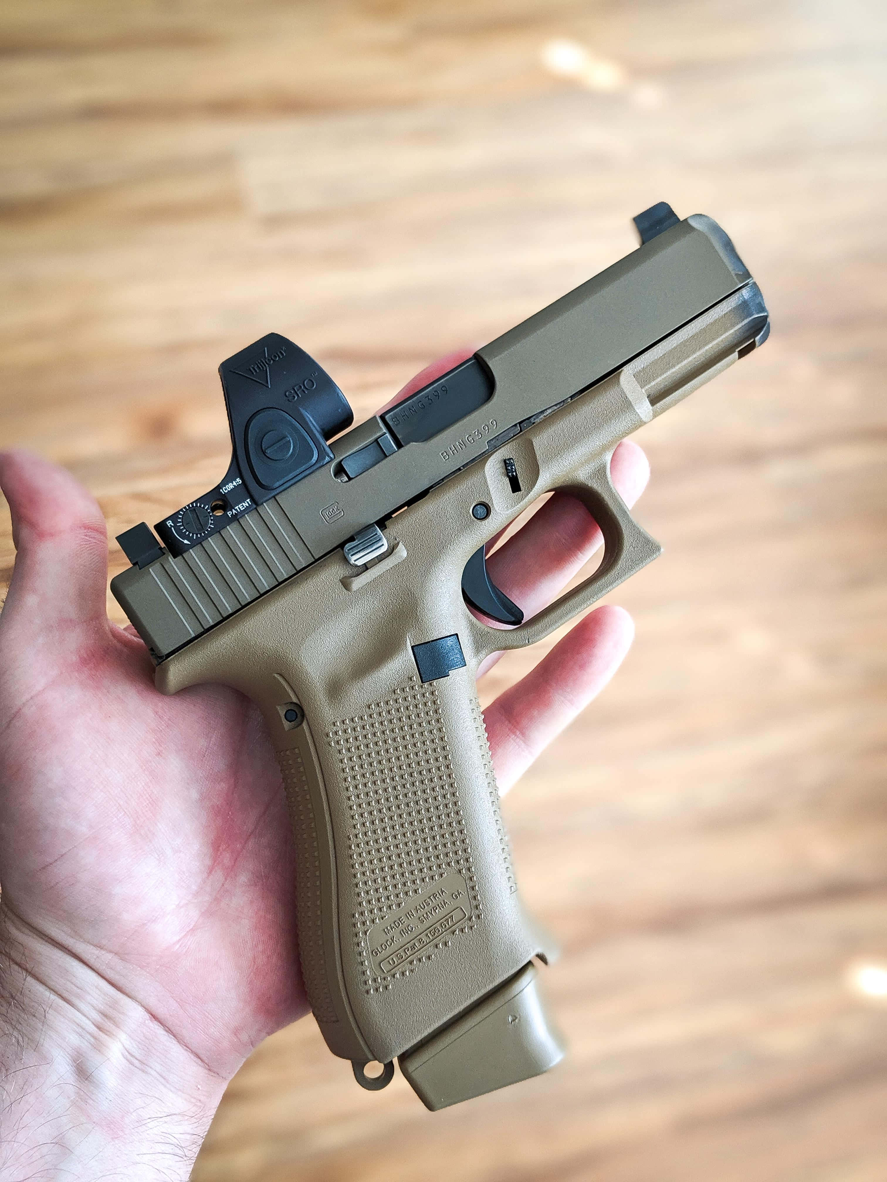 Glock 19 桌布 壁紙 — 一隻手拿著一把棕褐色格洛克手槍，配有紅點瞄準器，背景是模糊的木地板。