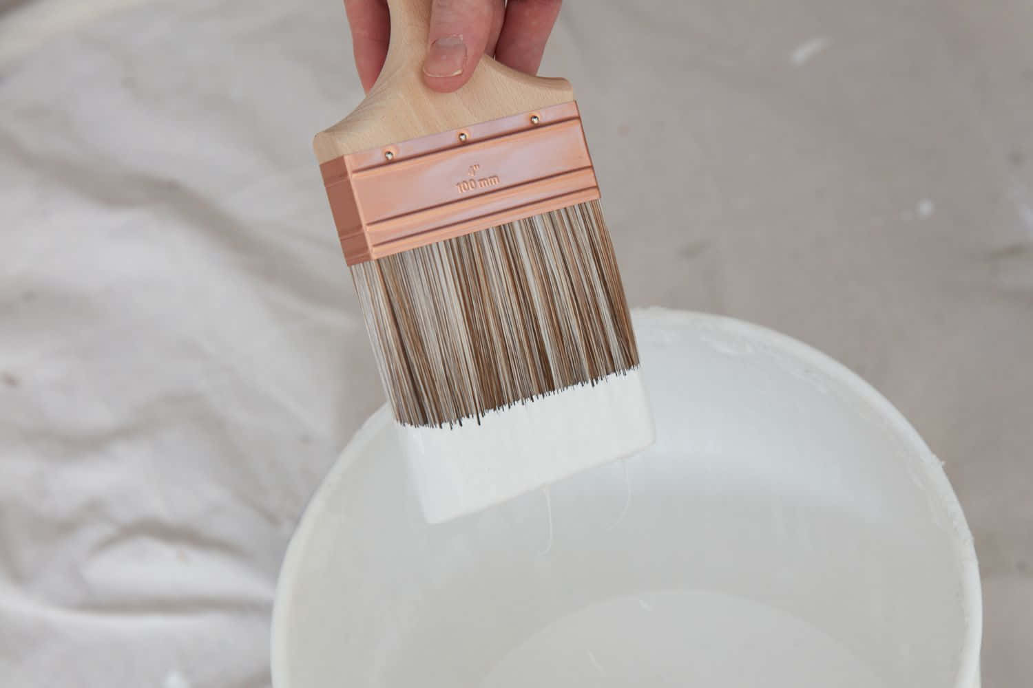 Paint Brush 桌布 壁紙 — 一隻手拿著一支100毫米油漆刷，白色油漆滴入一個白色桶中，背景是皺巴巴的白色背景。
