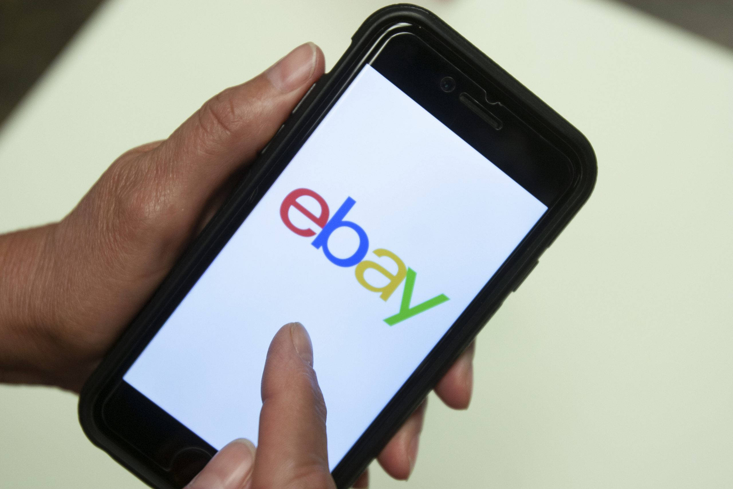 eBay 桌布 壁紙 — 一隻手拿著一支智慧型手機，在明亮的螢幕上顯示色彩繽紛的eBay標誌，背景為純色。