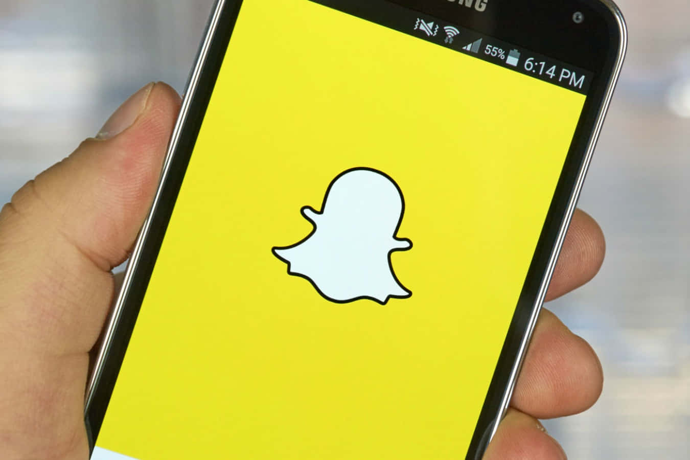 Snapchat 桌布 壁紙 — 一隻手拿著一支智慧型手機，螢幕上顯示Snapchat應用程式標誌，背景為亮黃色。