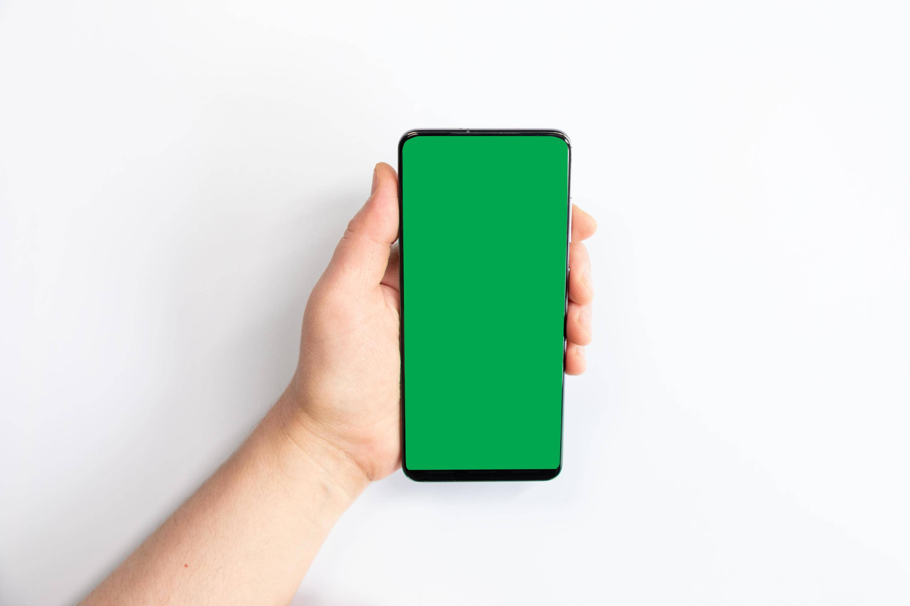 Green Screen 桌布 壁紙 — 一隻手拿著一支智慧型手機，螢幕為純綠色，背景是乾淨的白色。