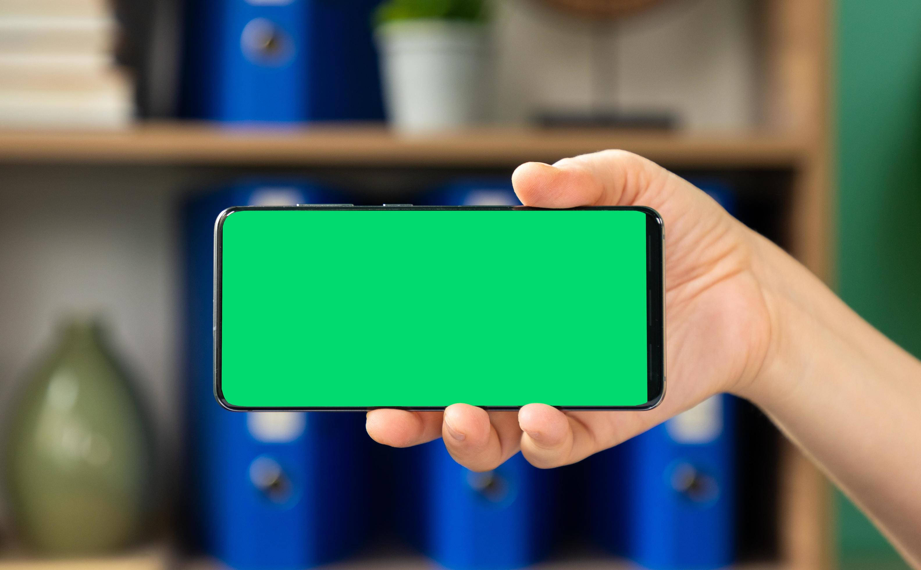 Green Minimalist 桌布 壁紙 — 一隻手拿著一支智慧型手機，螢幕為純綠色，背景是模糊的辦公室架子。