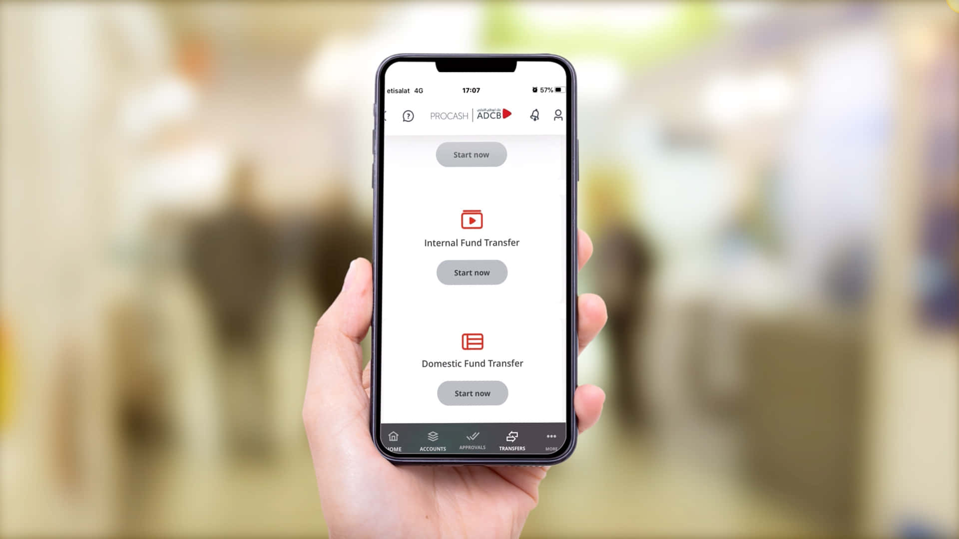 Mobile Banking 桌布 壁紙 — 一隻手拿著一支智慧型手機，顯示一個帶有轉帳選項的銀行應用程式，背景是模糊的室內環境。