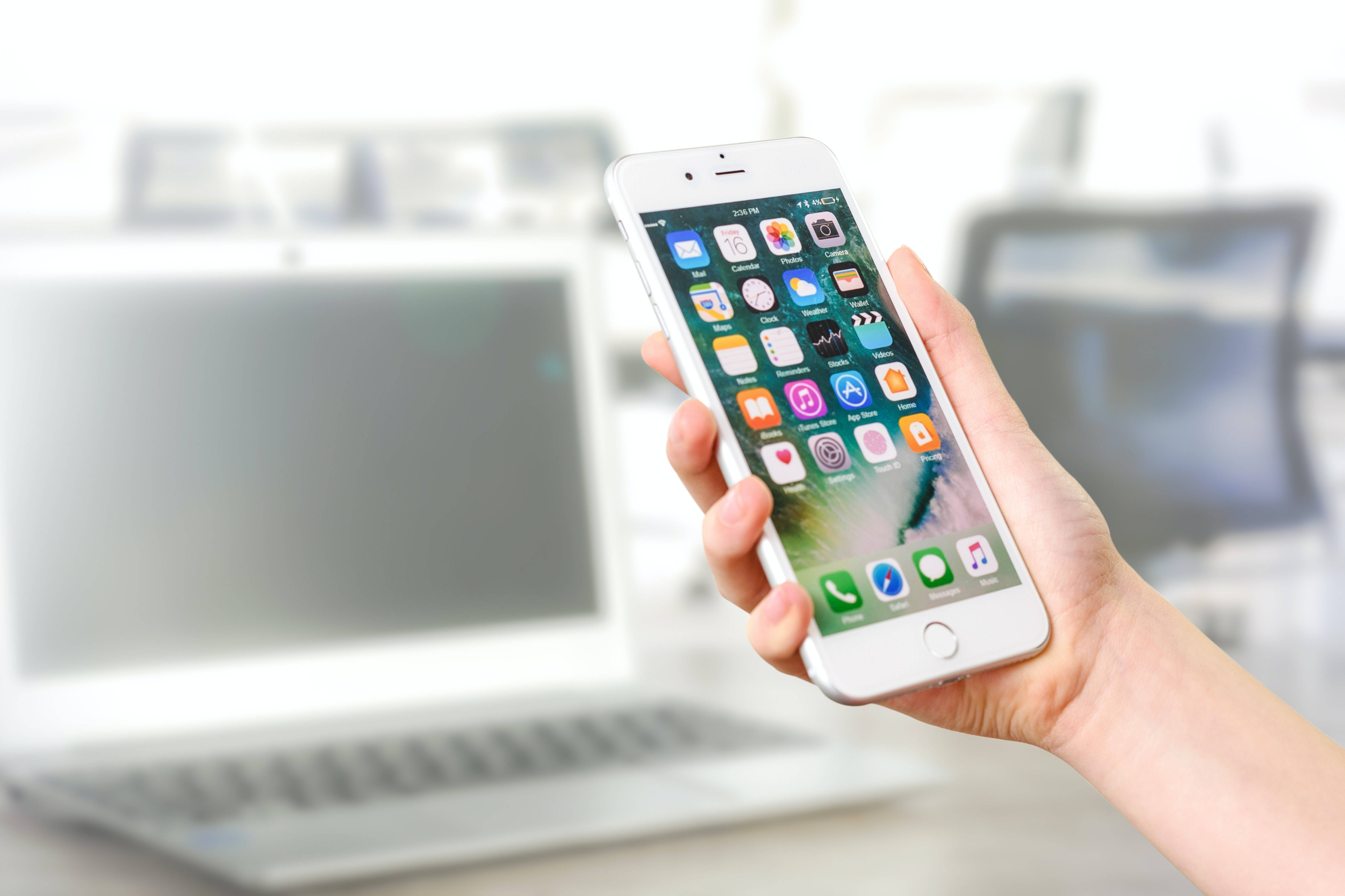 酷炫iPhone 桌布 壁紙 — 一隻手拿著一支白色智慧型手機，顯示色彩繽紛的應用程式圖示，背景有一台模糊的筆記型電腦。