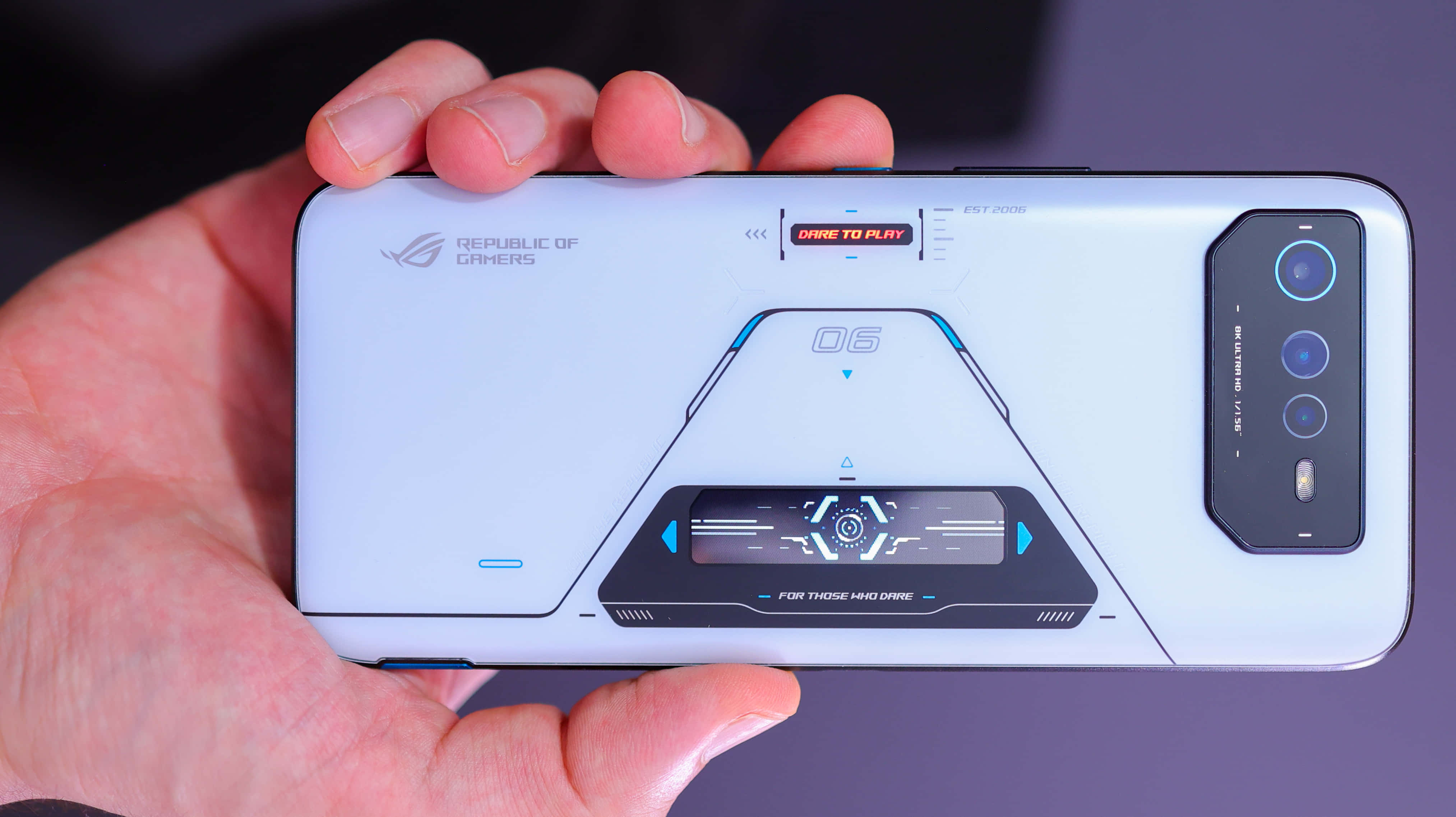 ASUS 桌布 壁紙 — 一隻手拿著一支白色遊戲手機，帶有藍色點綴和未來感相機模組，背景為紫色。