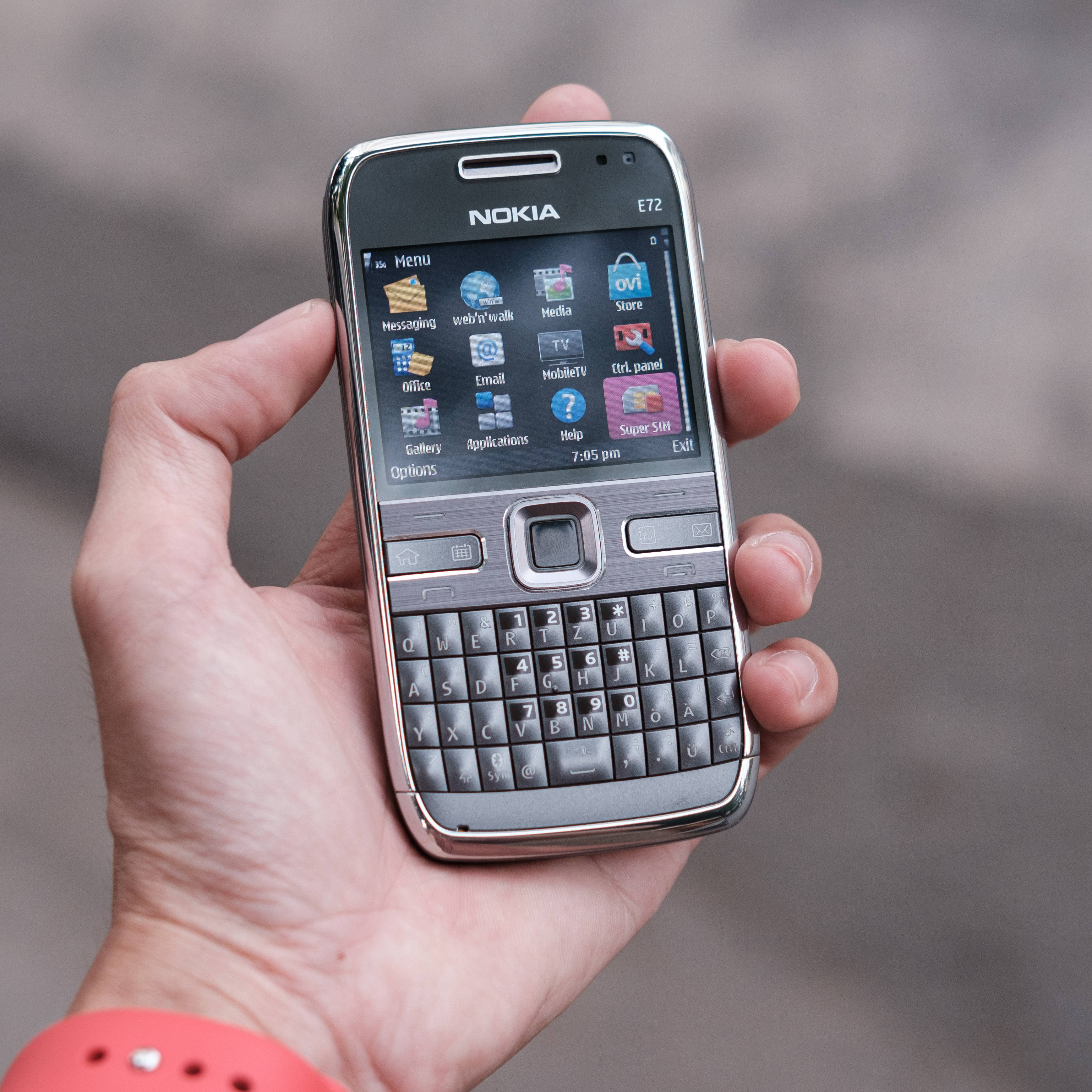 諾基亞 桌布 壁紙 — 一隻手拿著一支銀色Nokia E72智慧型手機，帶有實體QWERTY鍵盤和顯示應用程式及時間的活躍螢幕。