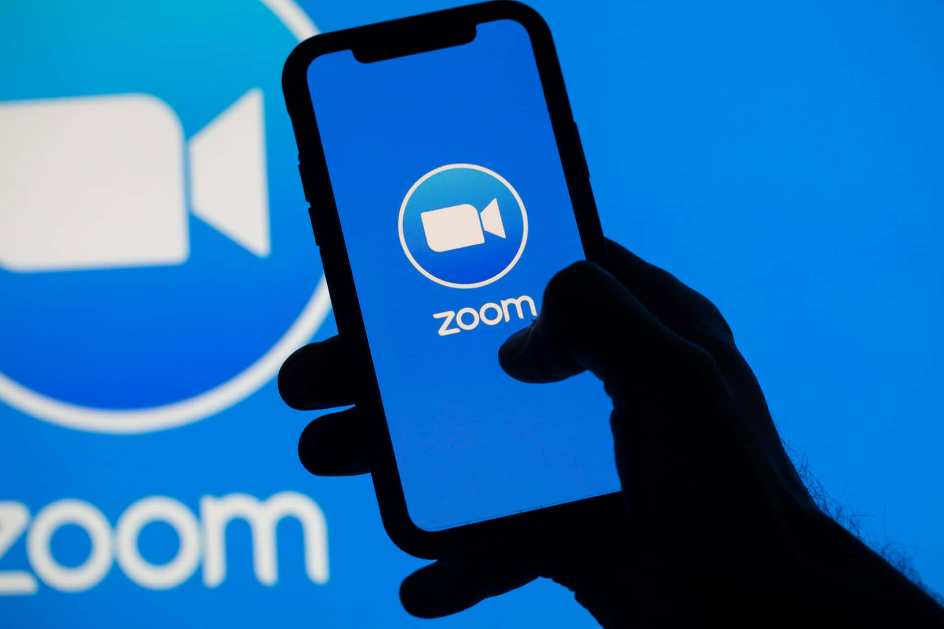 手拿著智慧型手機，開啟了Zoom應用程式，背景為藍色品牌背景。 — 放大 壁紙