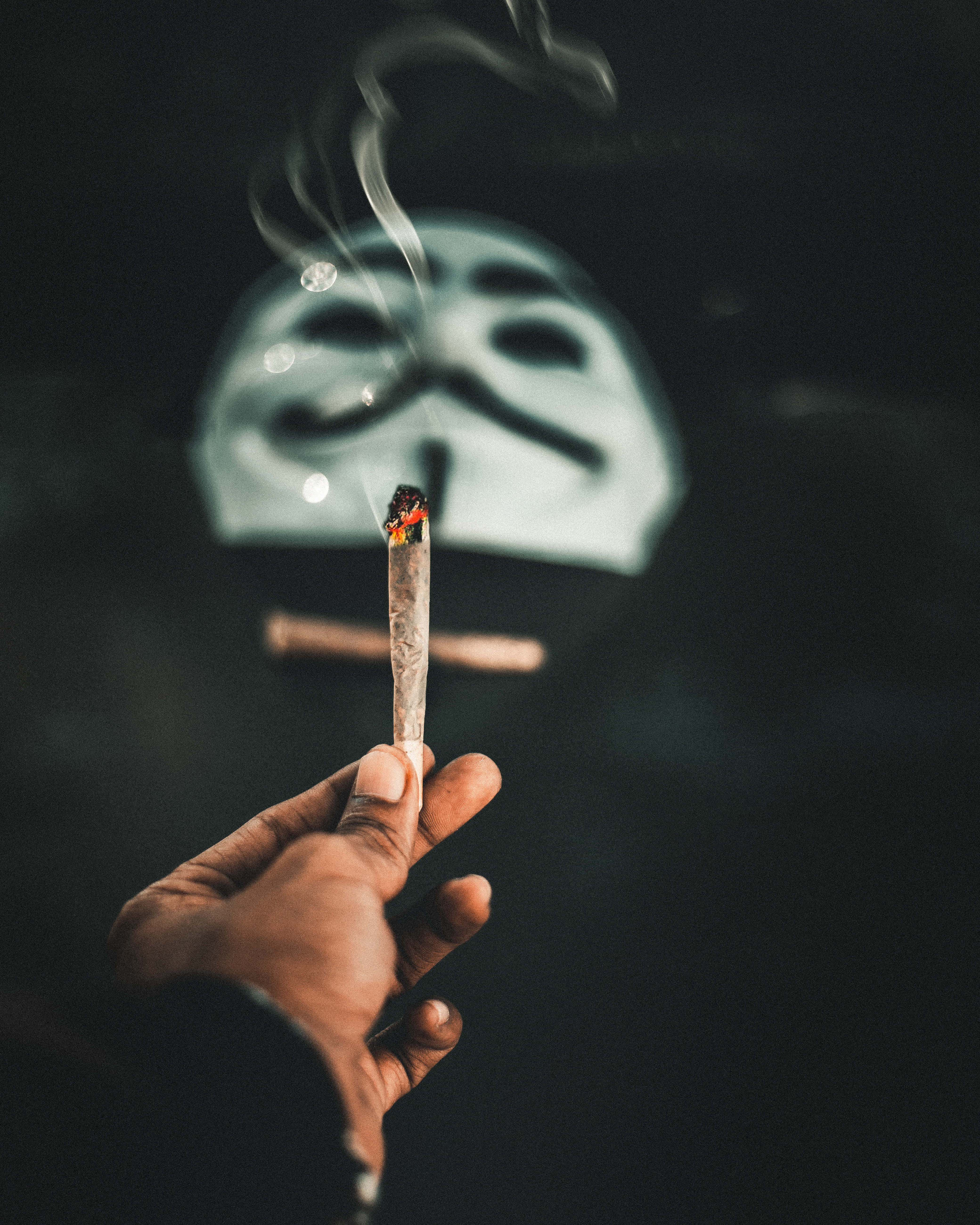 十字架 桌布 壁紙 — 一隻手拿著一支點燃的大麻煙，煙霧升起，背景是模糊的蓋伊·福克斯面具，背景黑暗。