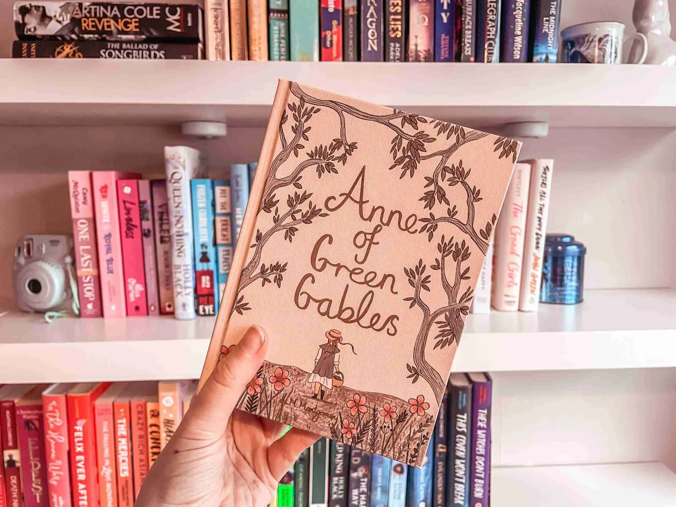 Aesthetic Books 桌布 壁紙 — 一隻手拿著一本名為《紅髮安妮》的書，書上有樹木和花朵插圖，背景是書架上色彩繽紛的書籍。