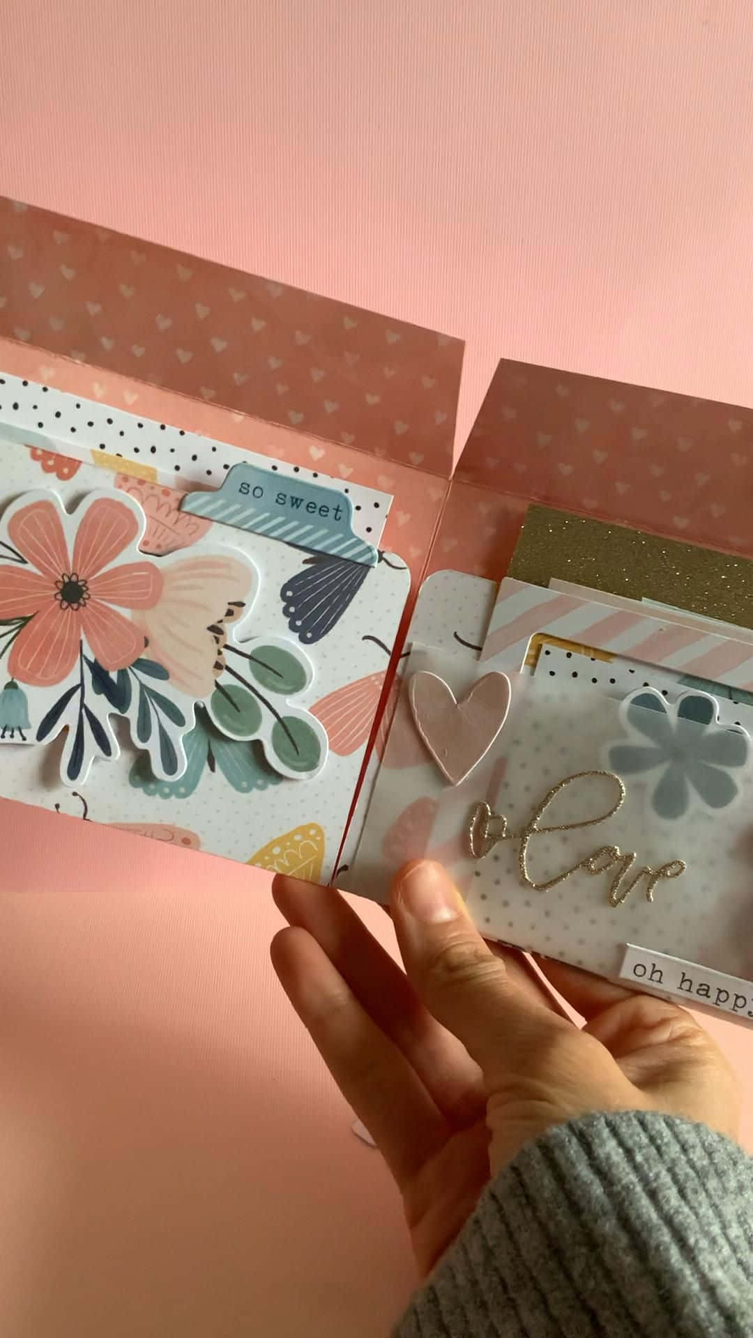 Scrapbooking 桌布 壁紙 — 一隻手拿著一本打開的手工剪貼簿，上面有花卉形狀的模切和閃亮的愛情標籤，背景是柔和的粉紅色。