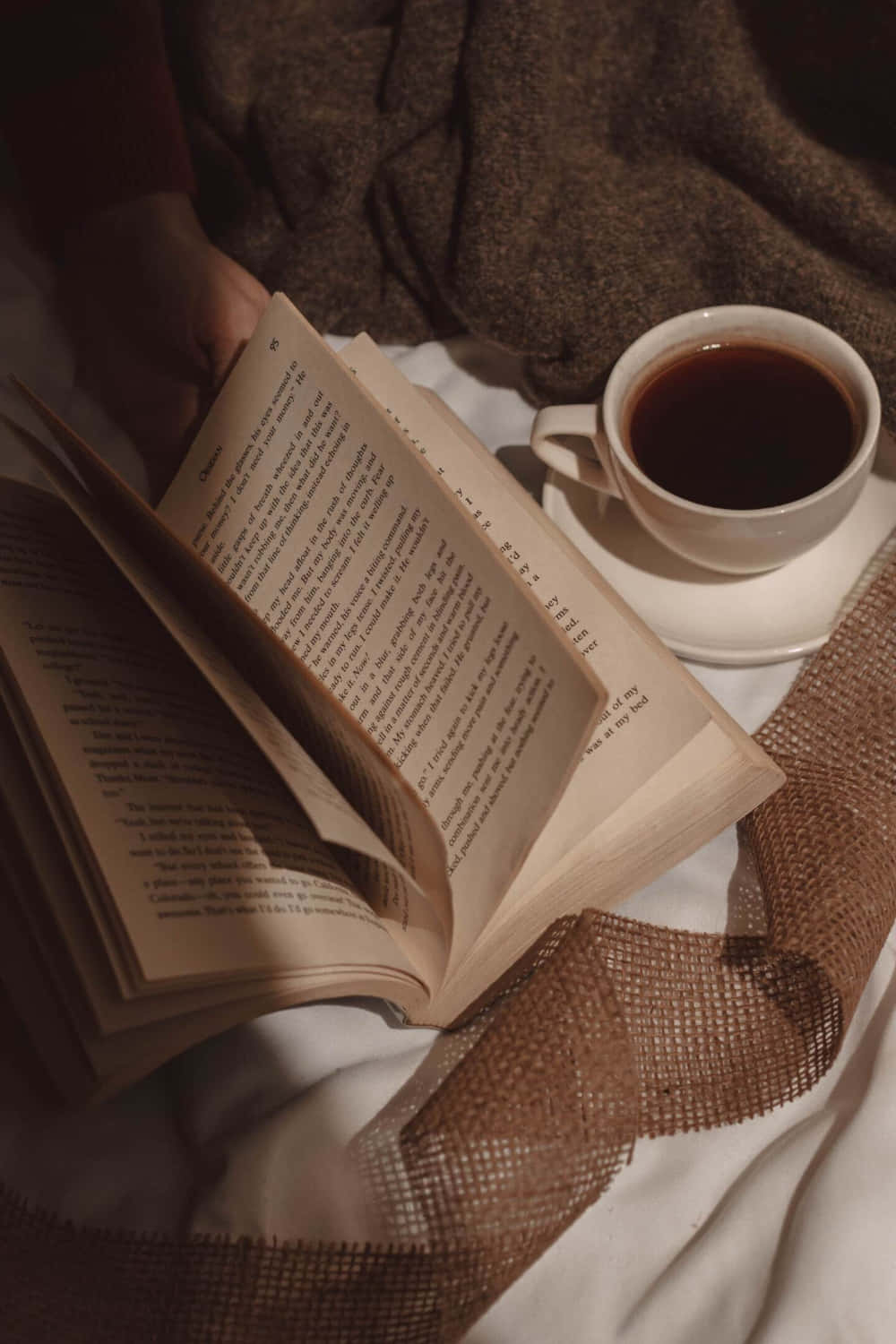 Aesthetic Book 桌布 壁紙 — 一隻手拿著一本打開的書，旁邊放著一杯咖啡，放在一張舒適的床上，床上有質感的棕色布料和白色床單。