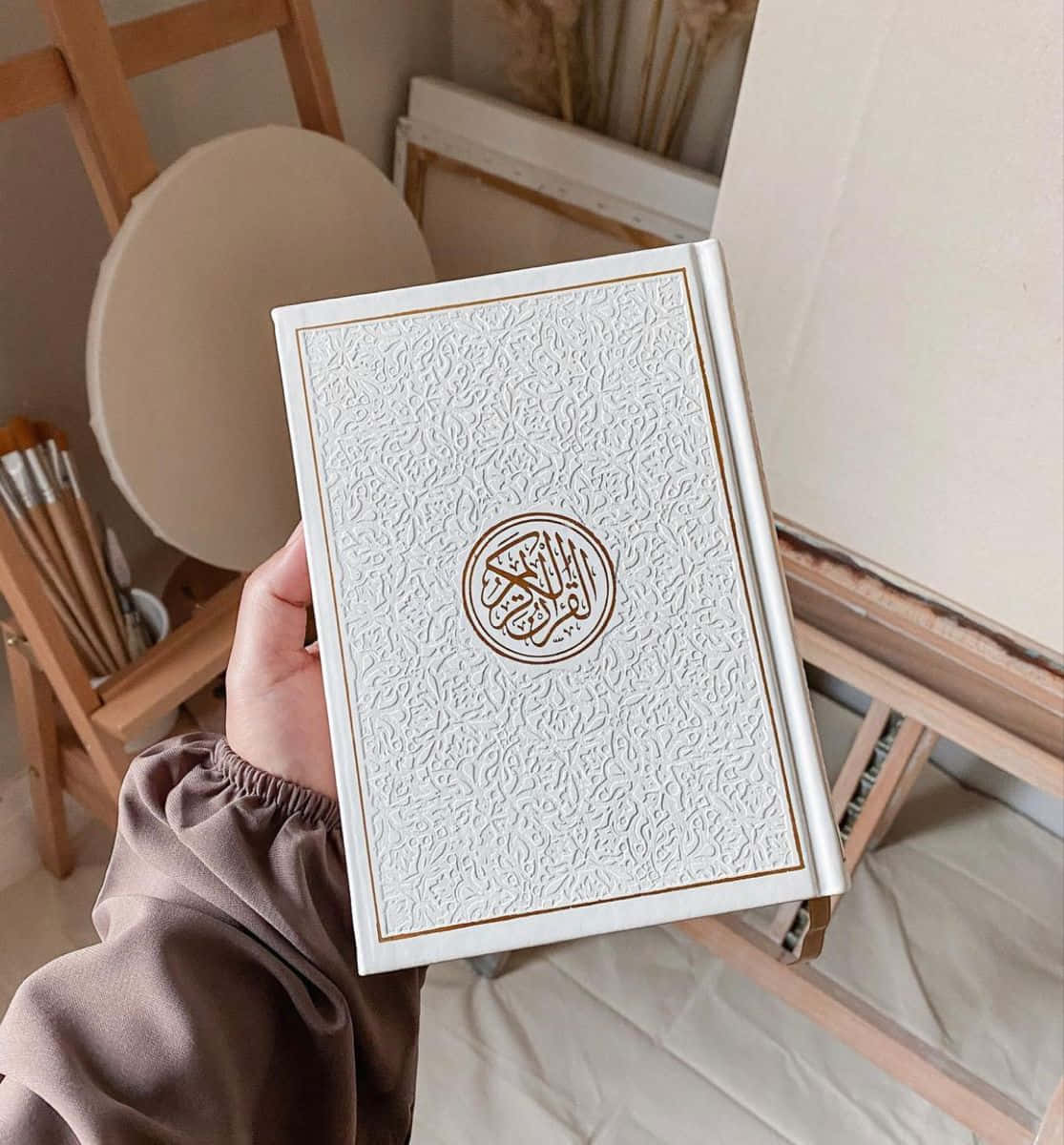 Quran 桌布 壁紙 — 一隻手拿著一本白色壓花古蘭經，帶有金色邊飾，背景是柔和的米色藝術家工作室。