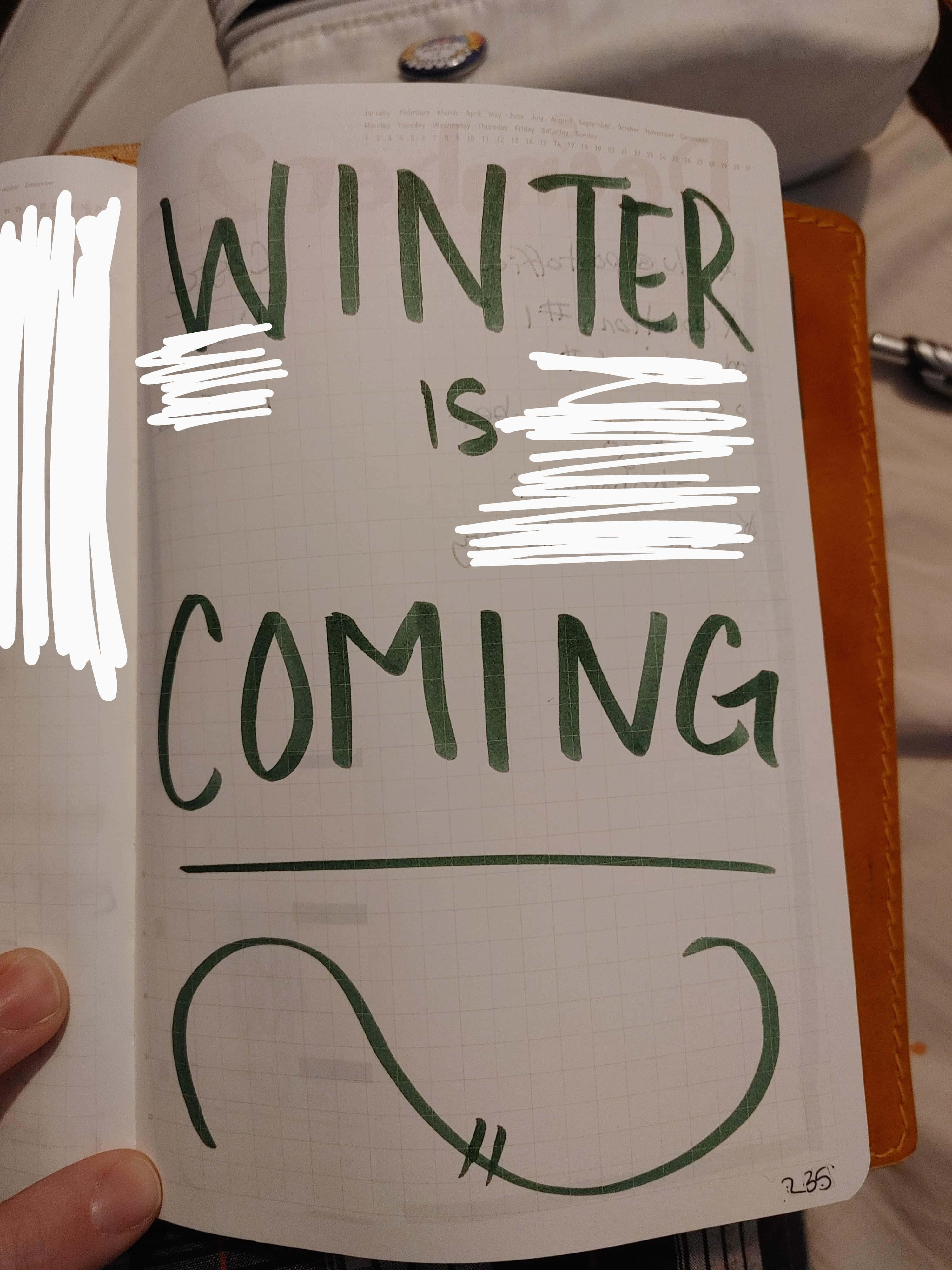 桌布 壁紙 — 一隻手拿著一本筆記本，上面用綠色墨水寫著「WINTER IS COMING」，部分被白色塗鴉遮蓋。